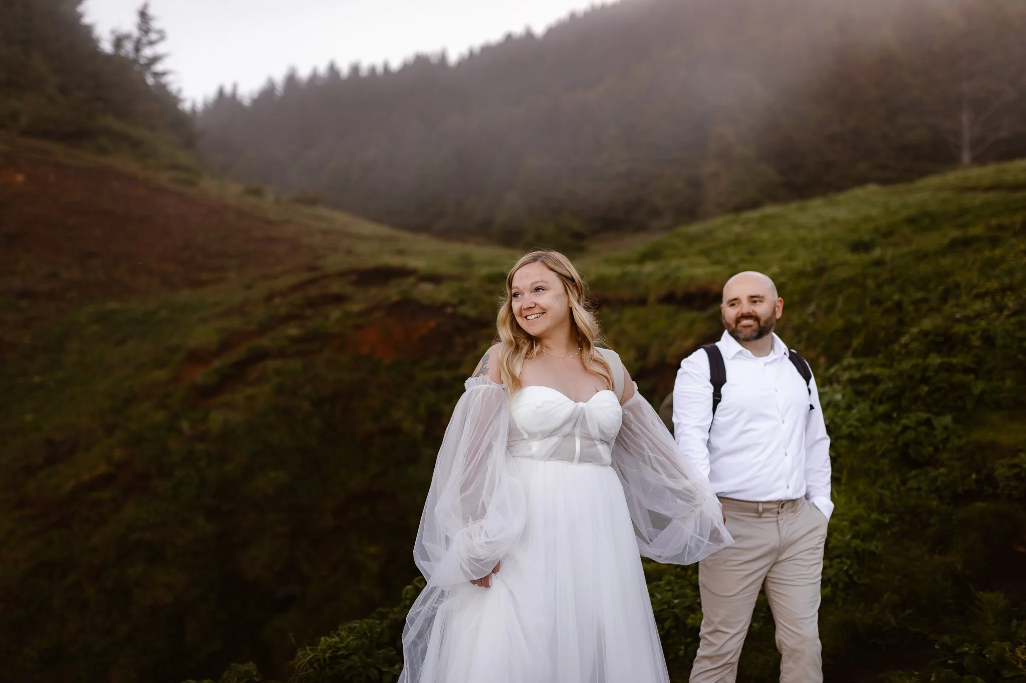 A couple, with the woman in a white wedding dress and the man in a white shirt, standing outdoors in front of a green hillside and trees.