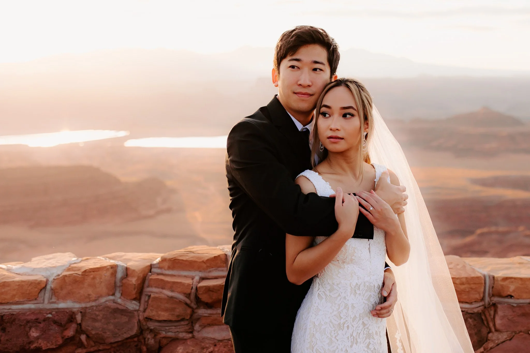A couple dressed in wedding attire standing outdoors near a brick wall, with scenic mountains and a body of water in the background during sunset.