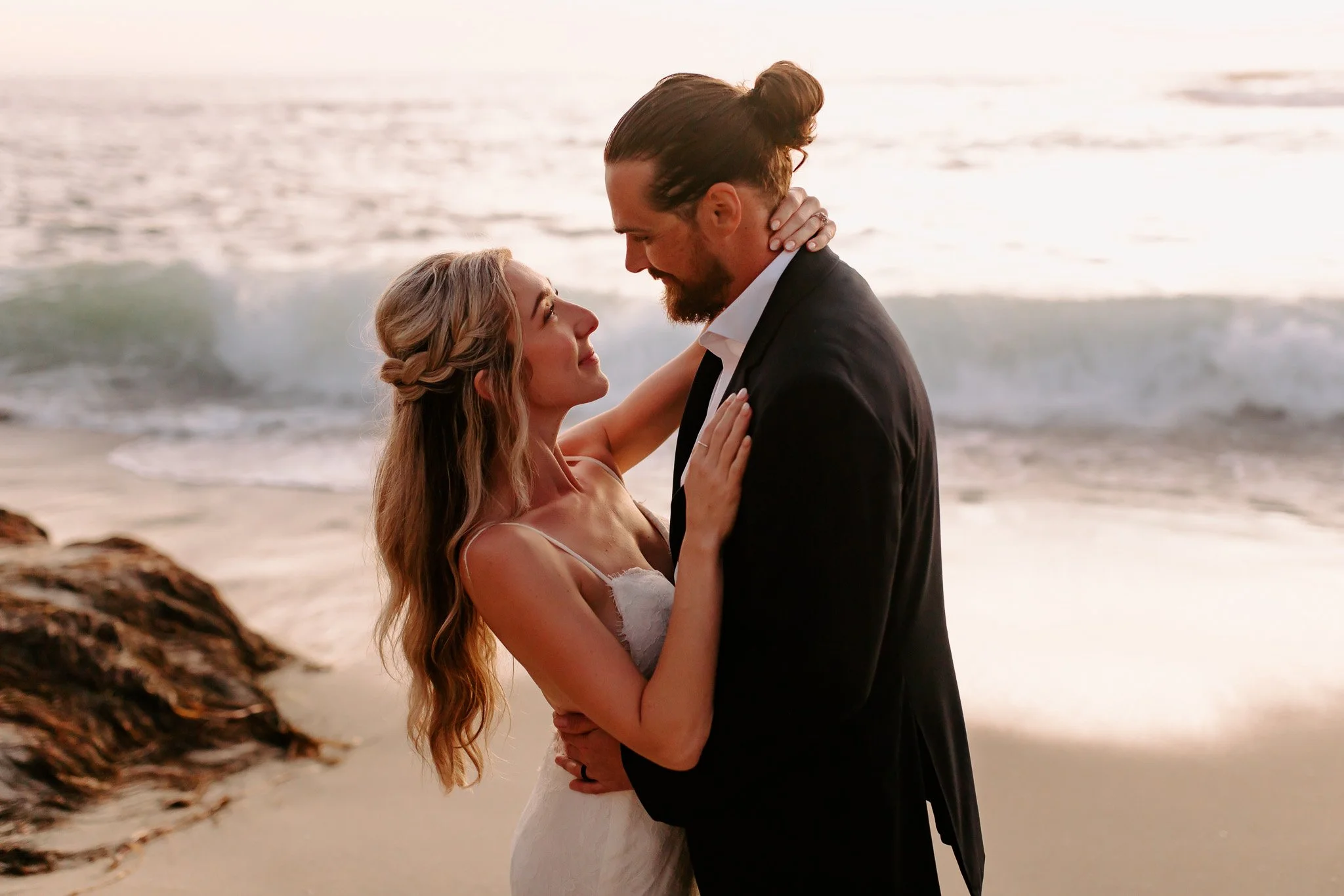 A couple stands close together at the beach, embracing each other with the ocean in the background during sunset.