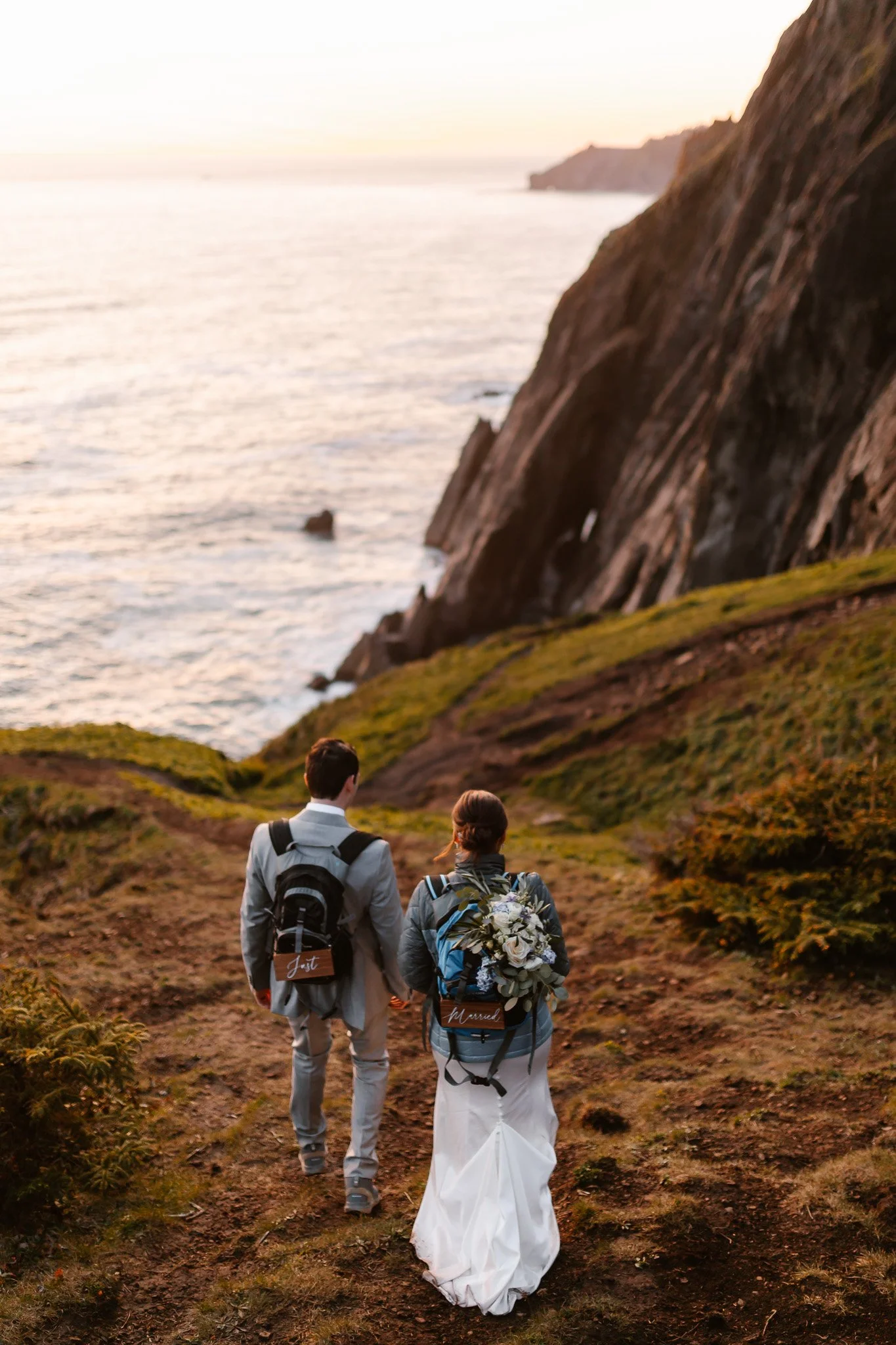 A bride and groom walking along a dirt trail near the coast with green moss-covered ground and cliffs in the background during sunset.