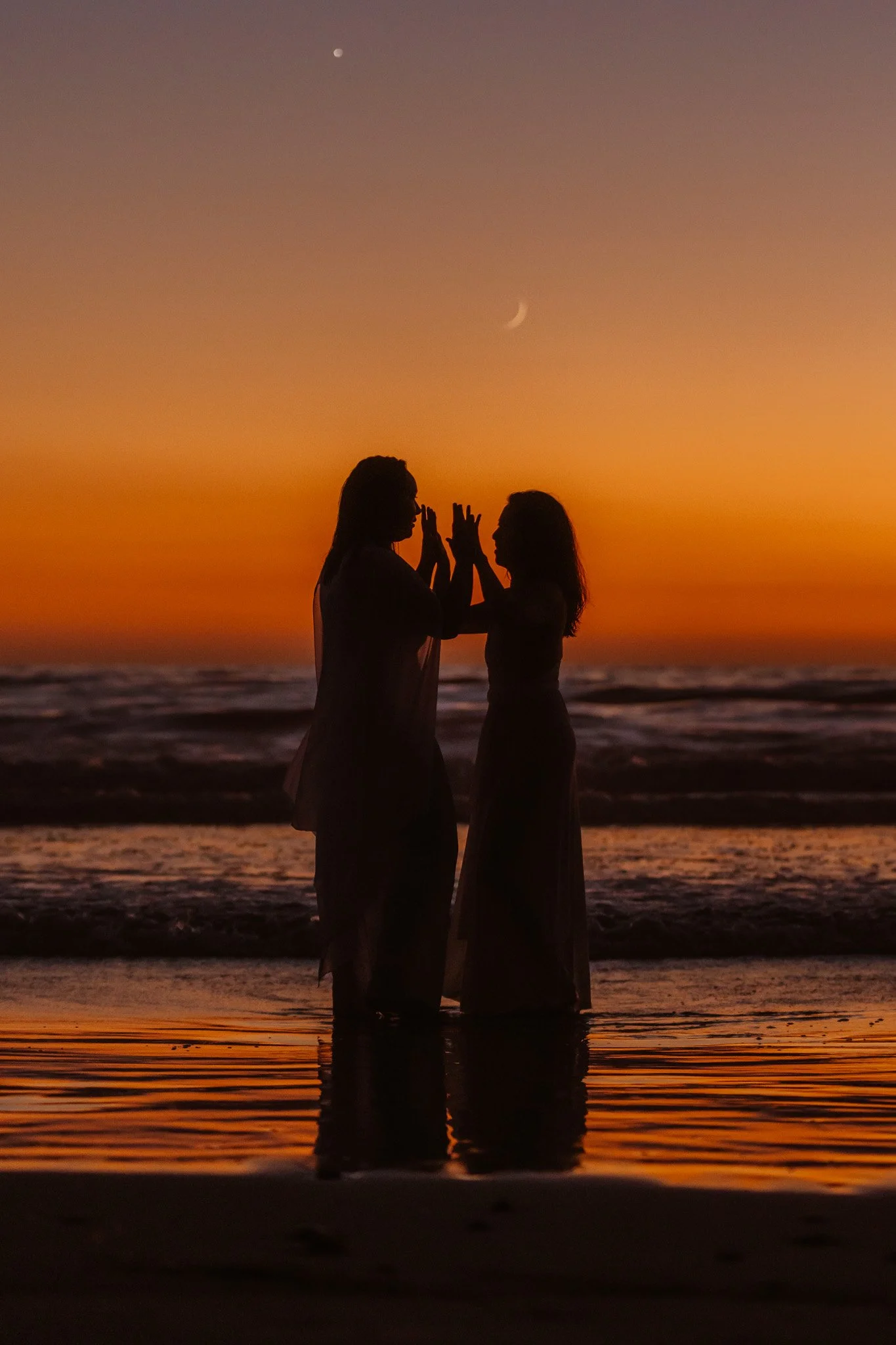 Silhouettes of two people standing in shallow water on a beach at sunset, with a crescent moon and bright star visible in the sky.