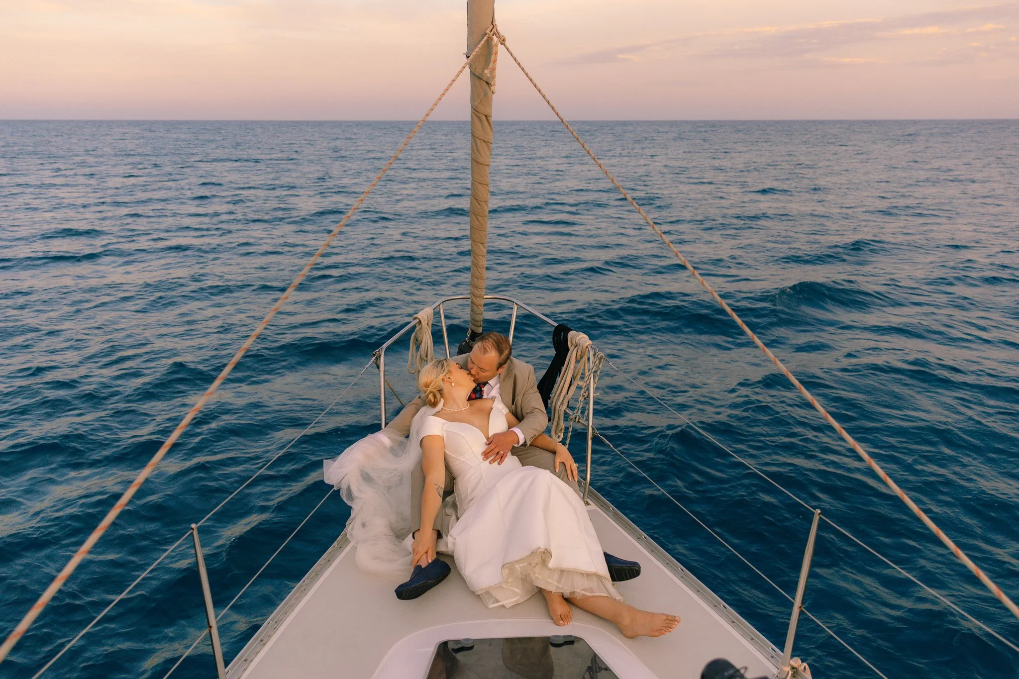 A wedding couple kissing and cuddling on the bow of a boat floating on the ocean at sunset with a pastel-colored sky.