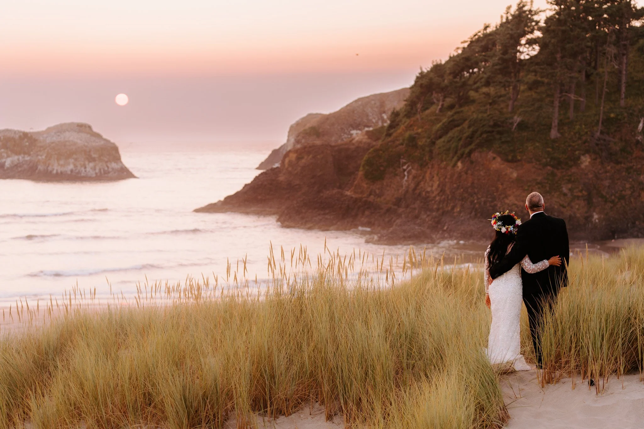 A bride and groom standing on a beach at sunset, embracing, with the bride wearing a white lace wedding gown and a floral crown, and the groom in a black suit, overlooking the ocean and rocky cliffs.