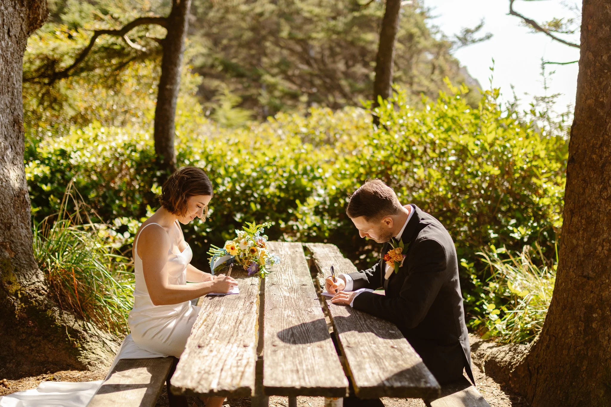A bride and groom sit on a wooden picnic table outdoors surrounded by trees and greenery, signing wedding documents during their wedding ceremony.