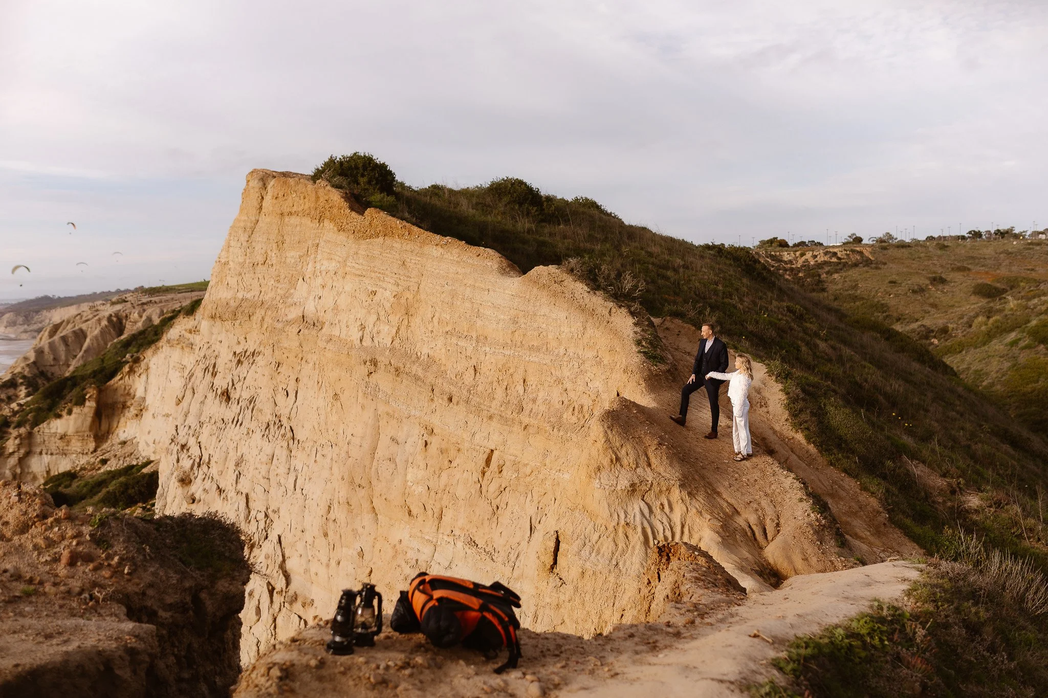 A man and a young girl holding hands walking along a rocky cliffside trail with panoramic views of a rugged landscape and cloudy sky.