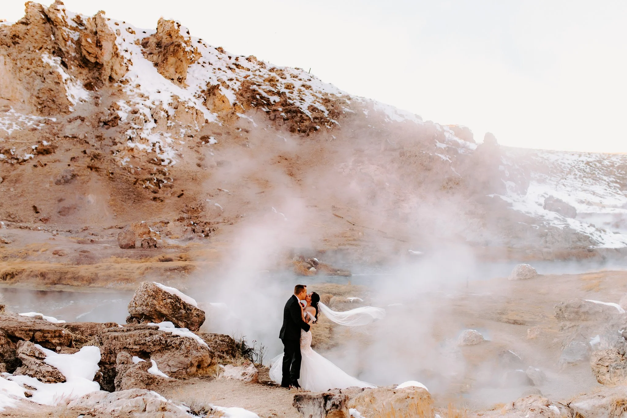 A bride and groom kissing near a steaming geothermal hot spring in a rocky, snow-dusted landscape with steam rising and a mountainous background.