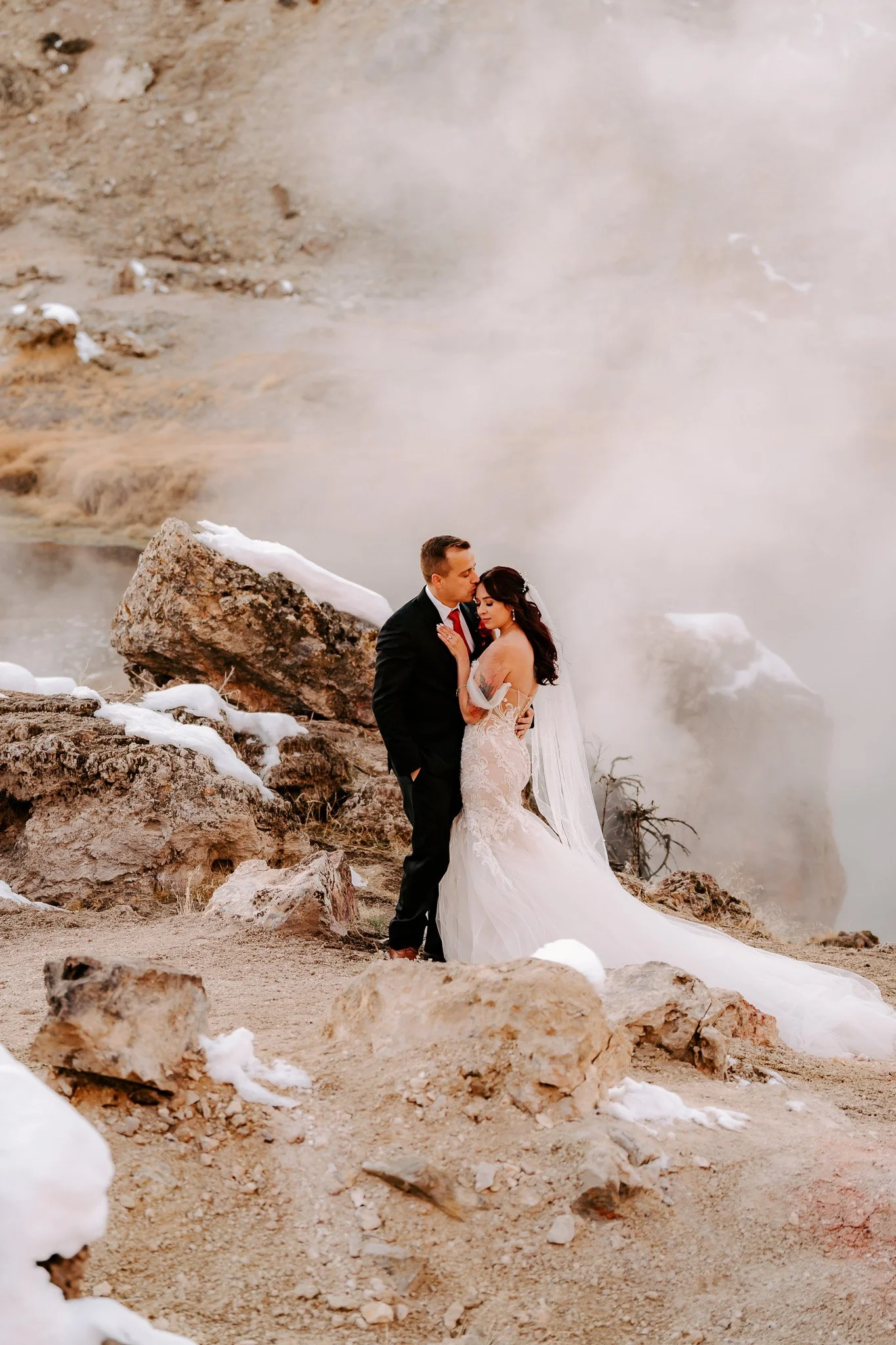 A bride and groom standing close together on rocky terrain with snow patches, surrounded by steam or fog rising from geothermal activity in the background.