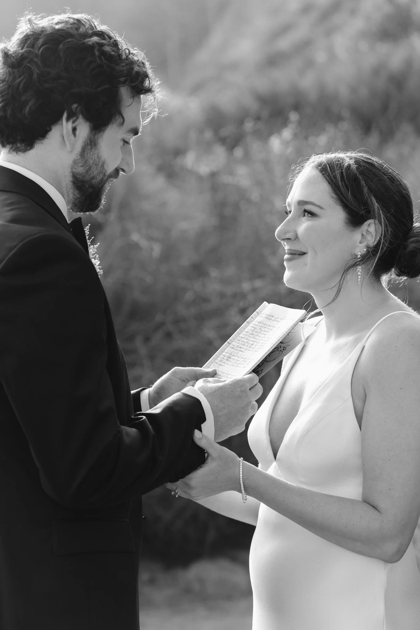 A black and white photo of a man in a tuxedo holding the hands of a pregnant woman in a wedding dress, both looking at each other, outdoors with blurred trees in the background.