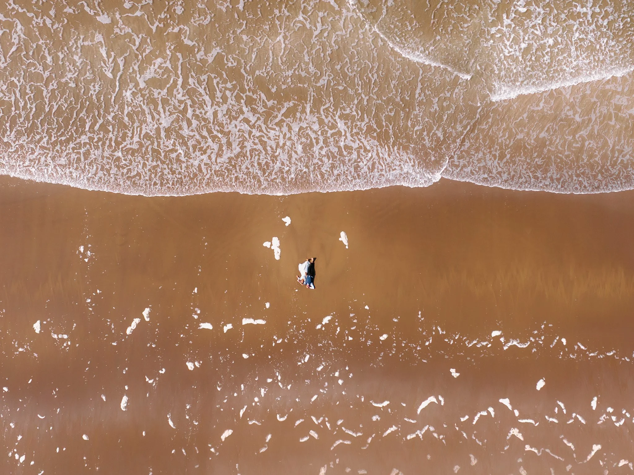Bird's eye view of two people lying on the beach near the shore, with the ocean waves approaching.