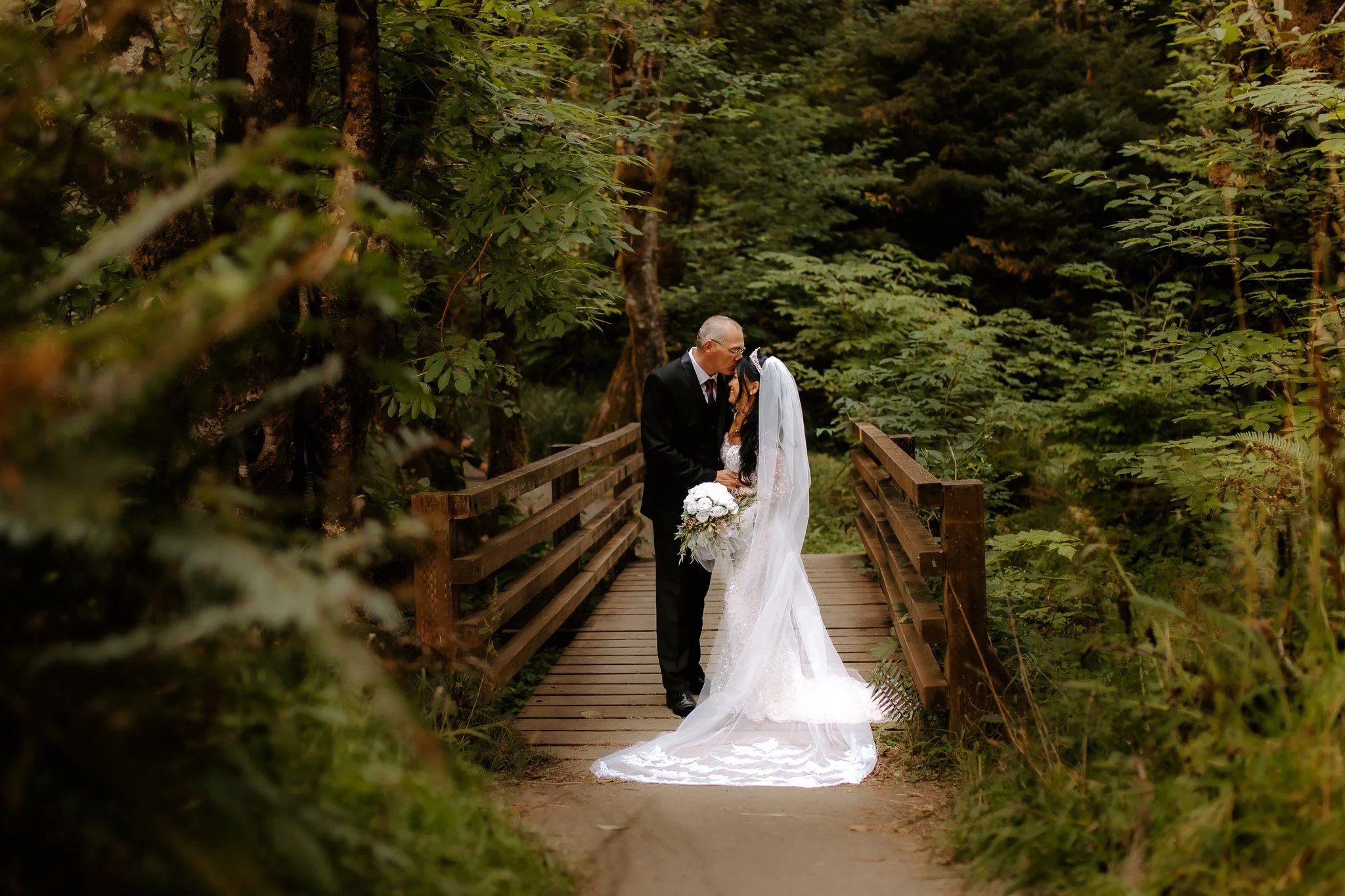 A bride and groom standing on a wooden bridge surrounded by lush green trees, sharing a tender moment.