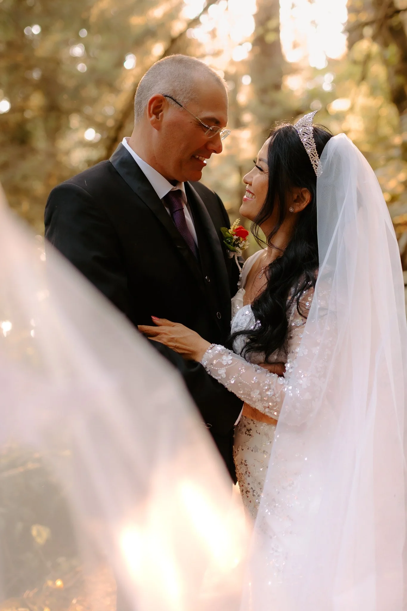 A bride and groom smiling and looking into each other's eyes during their wedding ceremony outdoors. The bride is wearing a white wedding dress and a veil, and the groom is dressed in a black suit with a boutonniere. Blurred trees and warm sunlight a