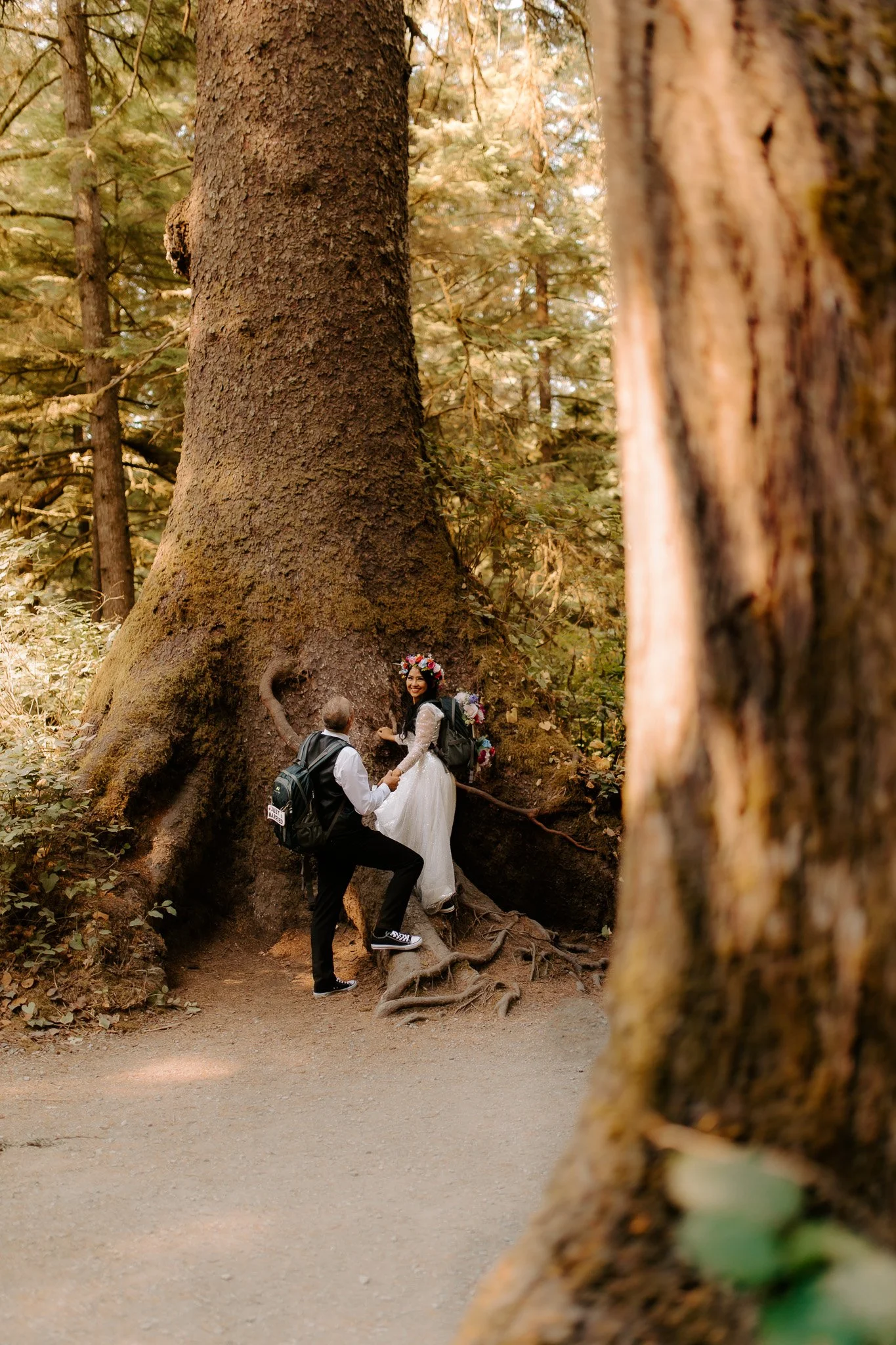 A couple in wedding attire holding hands and posing for a photo in a forest, with large trees and sunlight filtering through the leaves.
