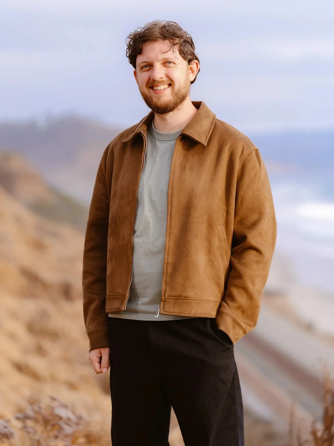 Smiling man standing outdoors on a beach during daytime, wearing a brown jacket and black pants.