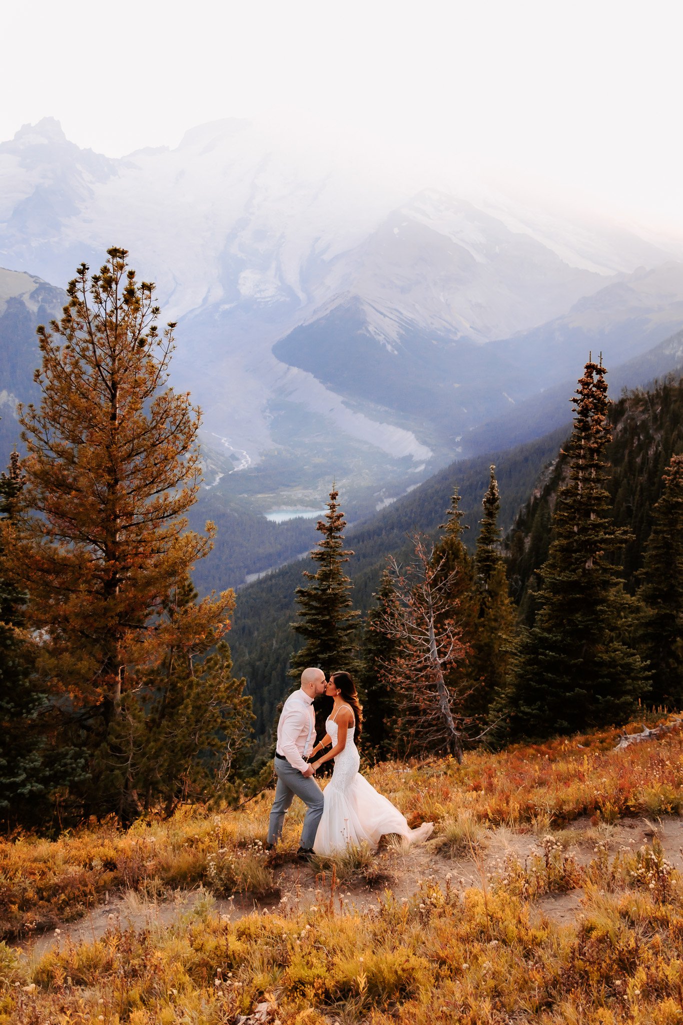 A wedding couple kissing in a mountain landscape with trees, mountains, and snow in the background.
