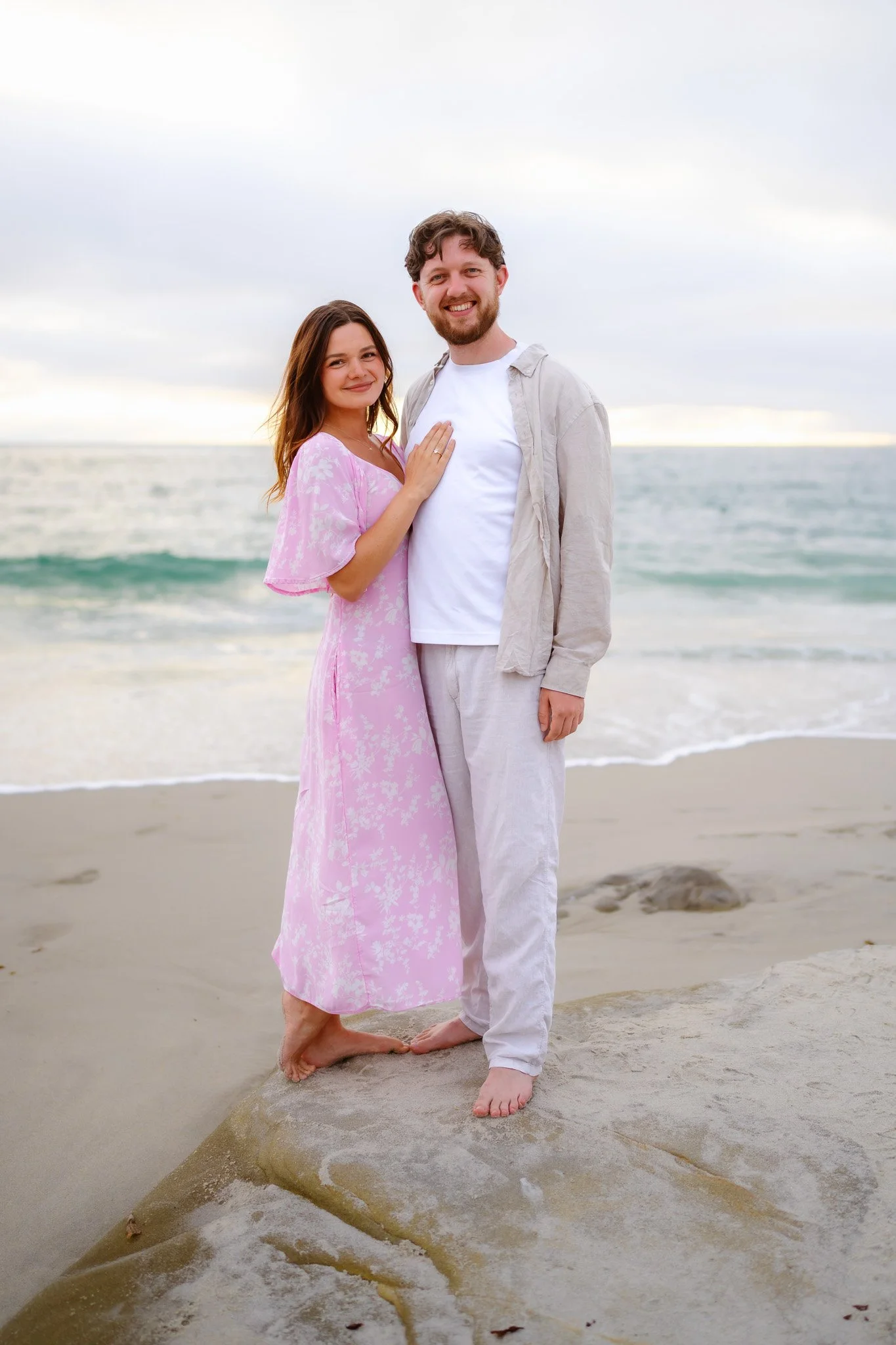 A smiling couple stands on a rock at the beach with ocean waves behind them. The woman wears a pink dress, and the man wears a white shirt and light pants. They are barefoot on the sand, and the sky is cloudy with a hint of sunset.