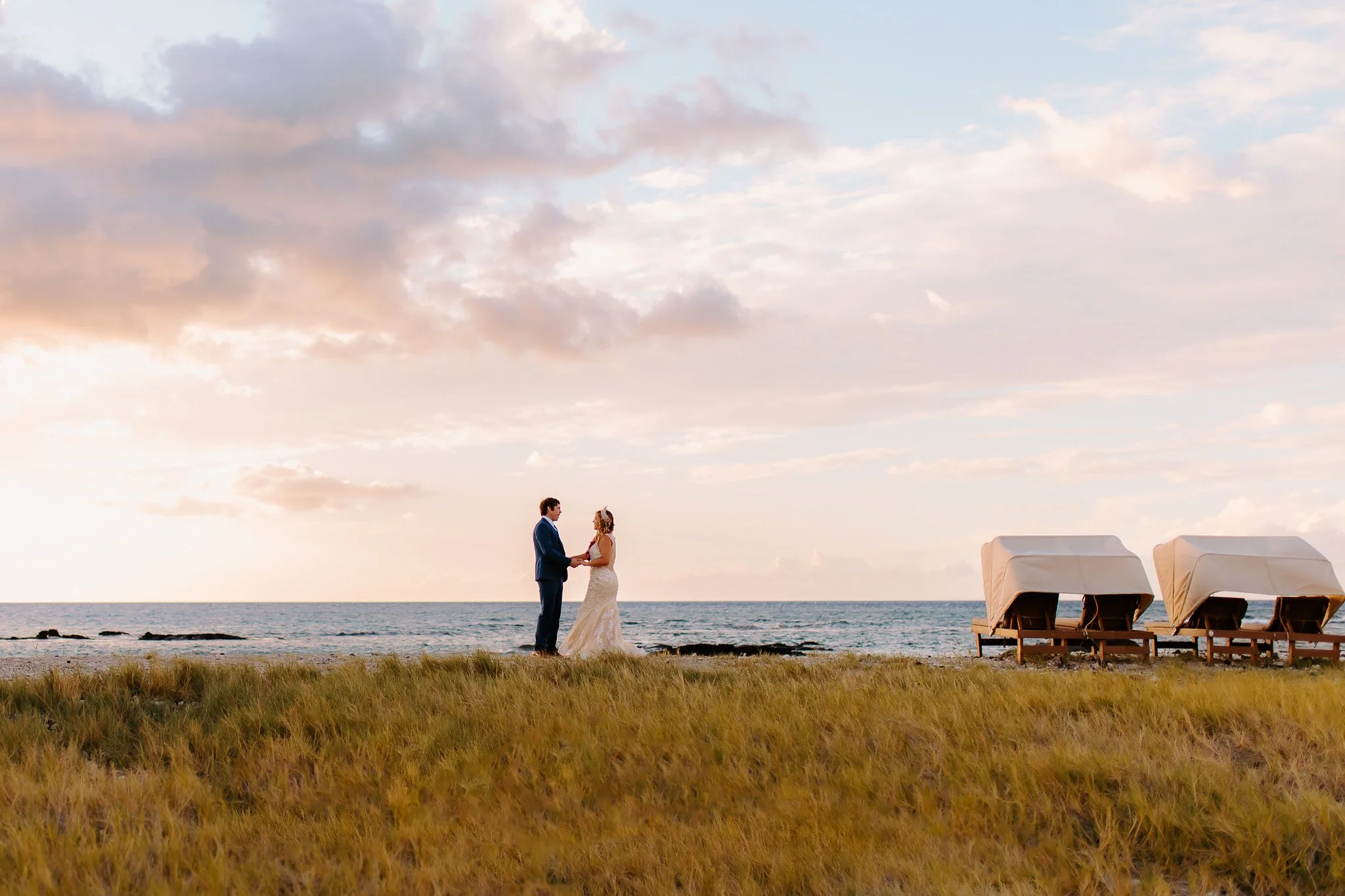 A couple holding hands on a beach during sunset, with the ocean in the background and two white lounge chairs to the right.