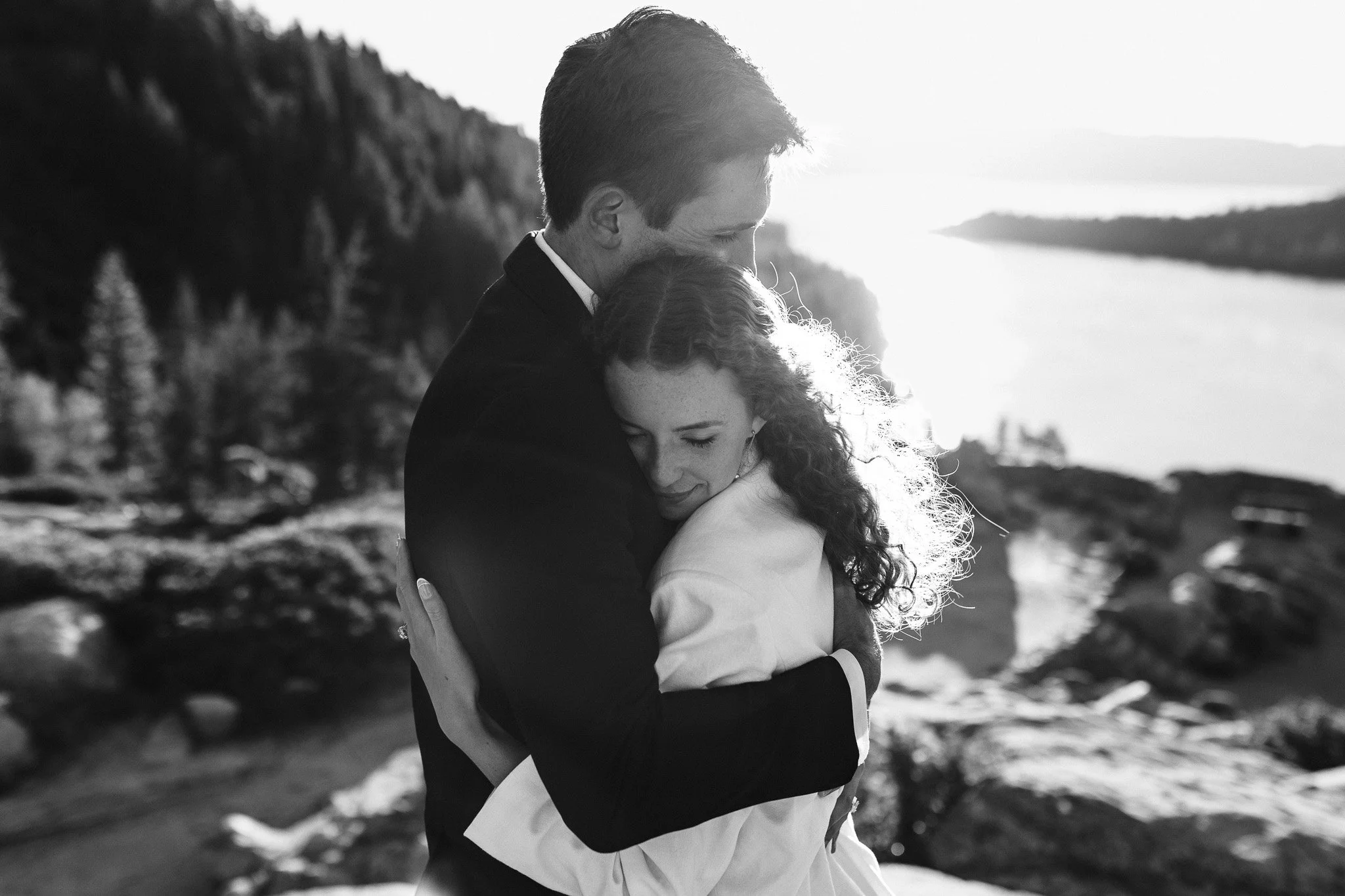 A black and white photo of a couple hugging outdoors on a rocky coastline with water and hills in the background. The man is dressed in a suit and the woman in a light dress. The man is leaning his head down to the woman's head, and both appear to be
