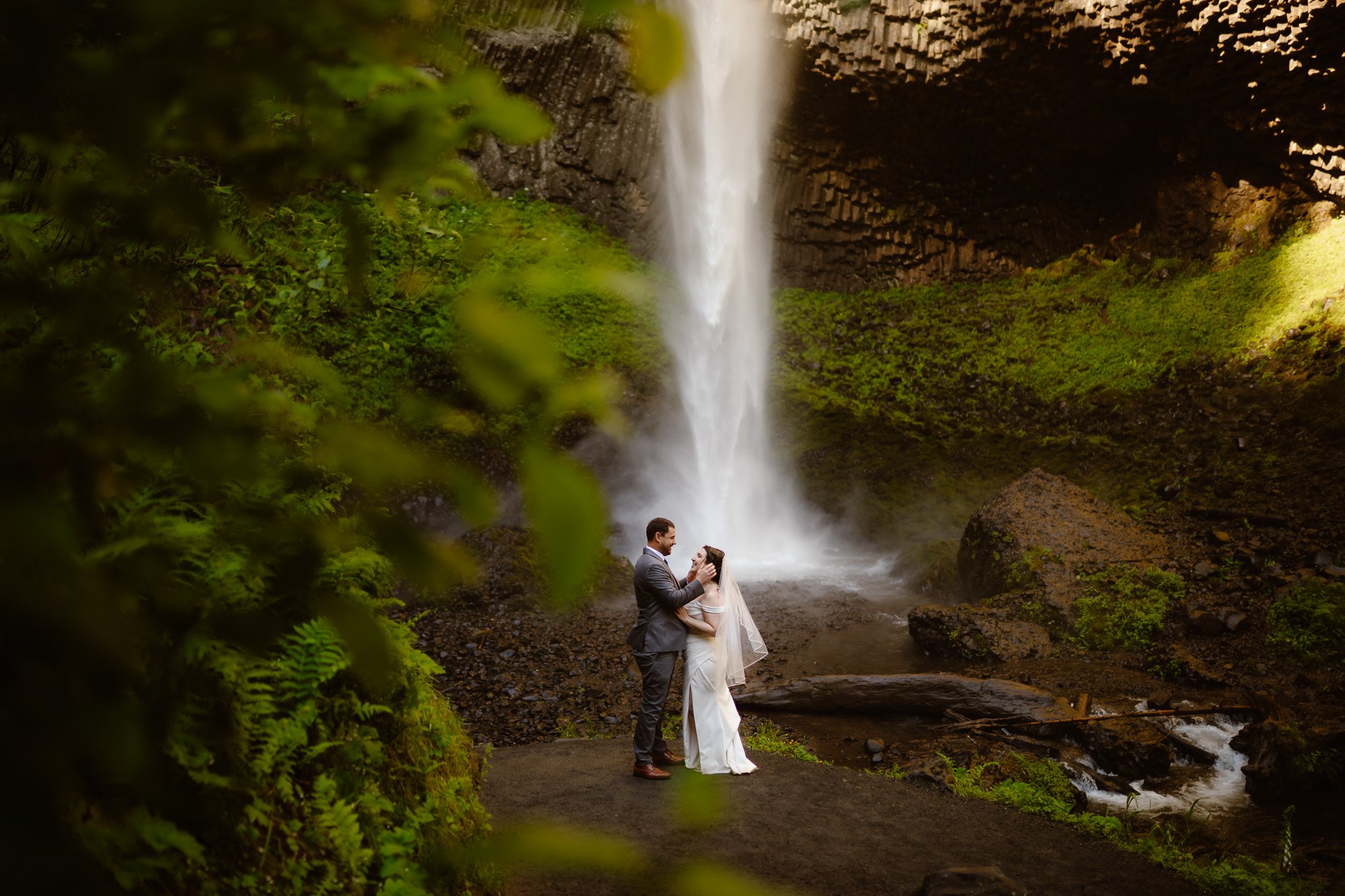 A couple dressed in wedding attire standing by a waterfall in a lush, green forest. The groom is in a suit, and the bride is in a white wedding dress with a veil. They are holding each other and gazing into each other's eyes.