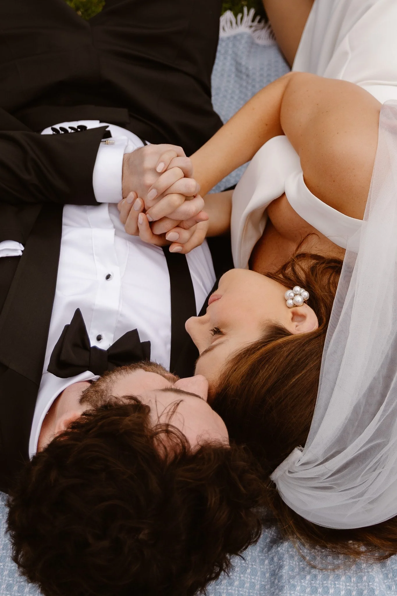A man and woman lying on the floor, holding hands, with their eyes closed, dressed in formal attire. The man is in a tuxedo with a bow tie, and the woman is wearing a white dress with a pearl earrings and a veil.