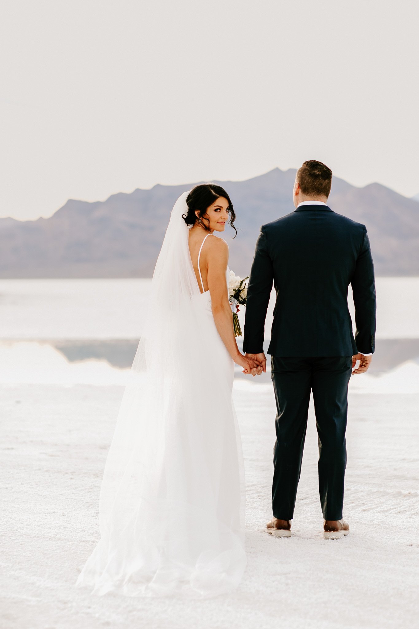 A bride and groom holding hands on a salt flat during their wedding, with mountains in the background and a clear, bright sky.