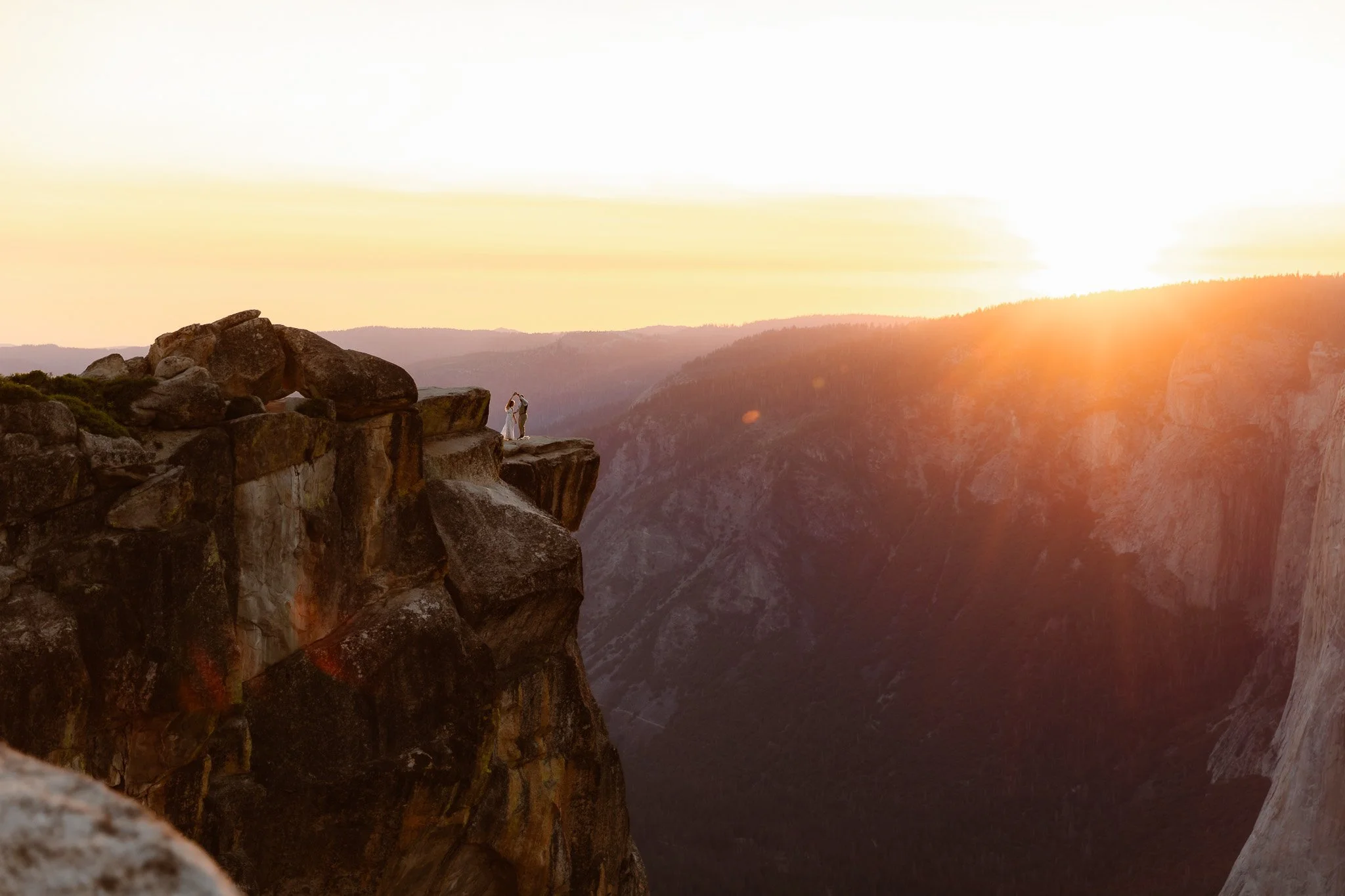A couple standing on a rock ledge at sunset, with mountains and valleys in the background.