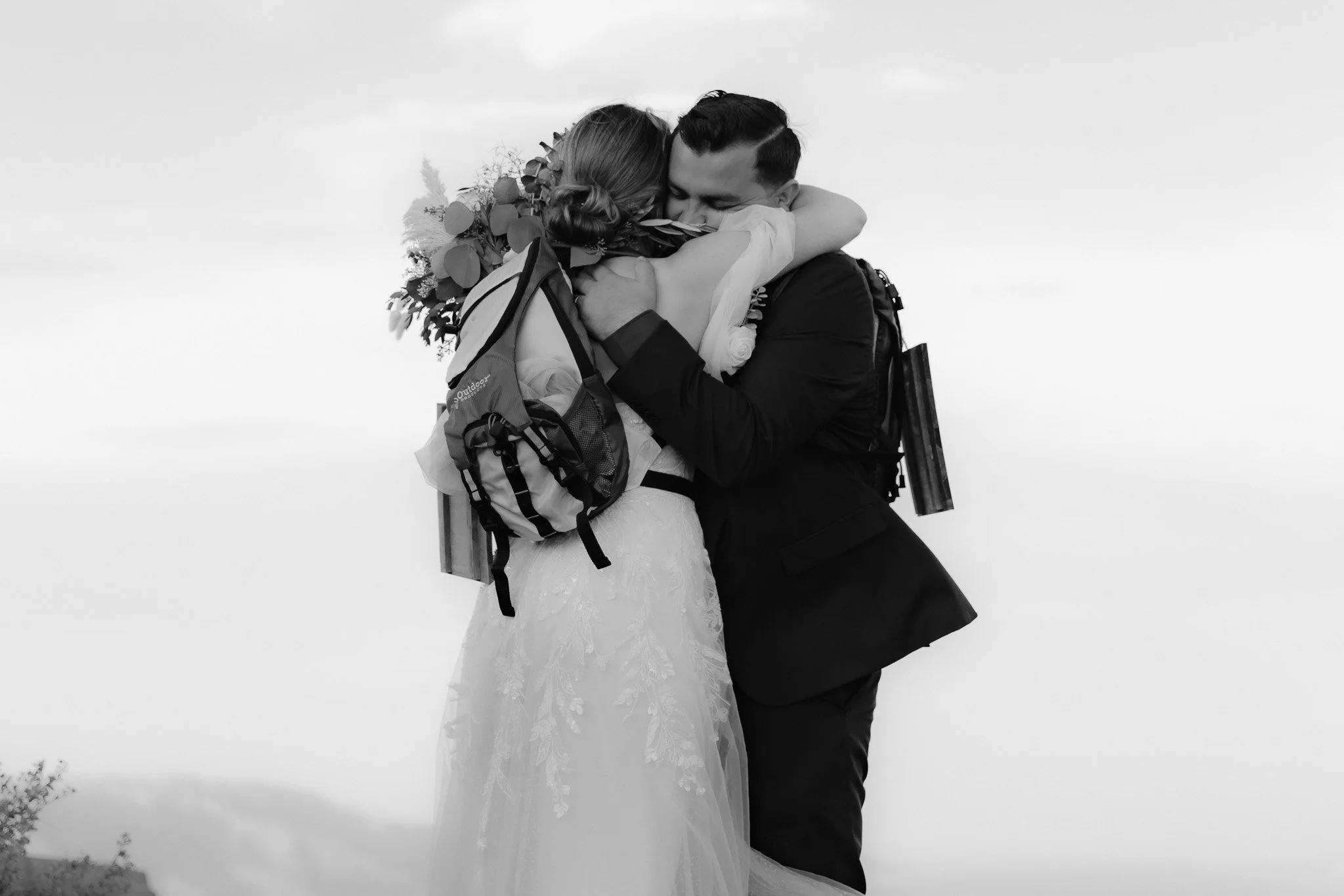 A black-and-white photo of a bride and groom hugging outdoors, with the bride wearing a wedding dress and holding a bouquet of flowers, and the groom in a suit. They are embracing each other warmly.