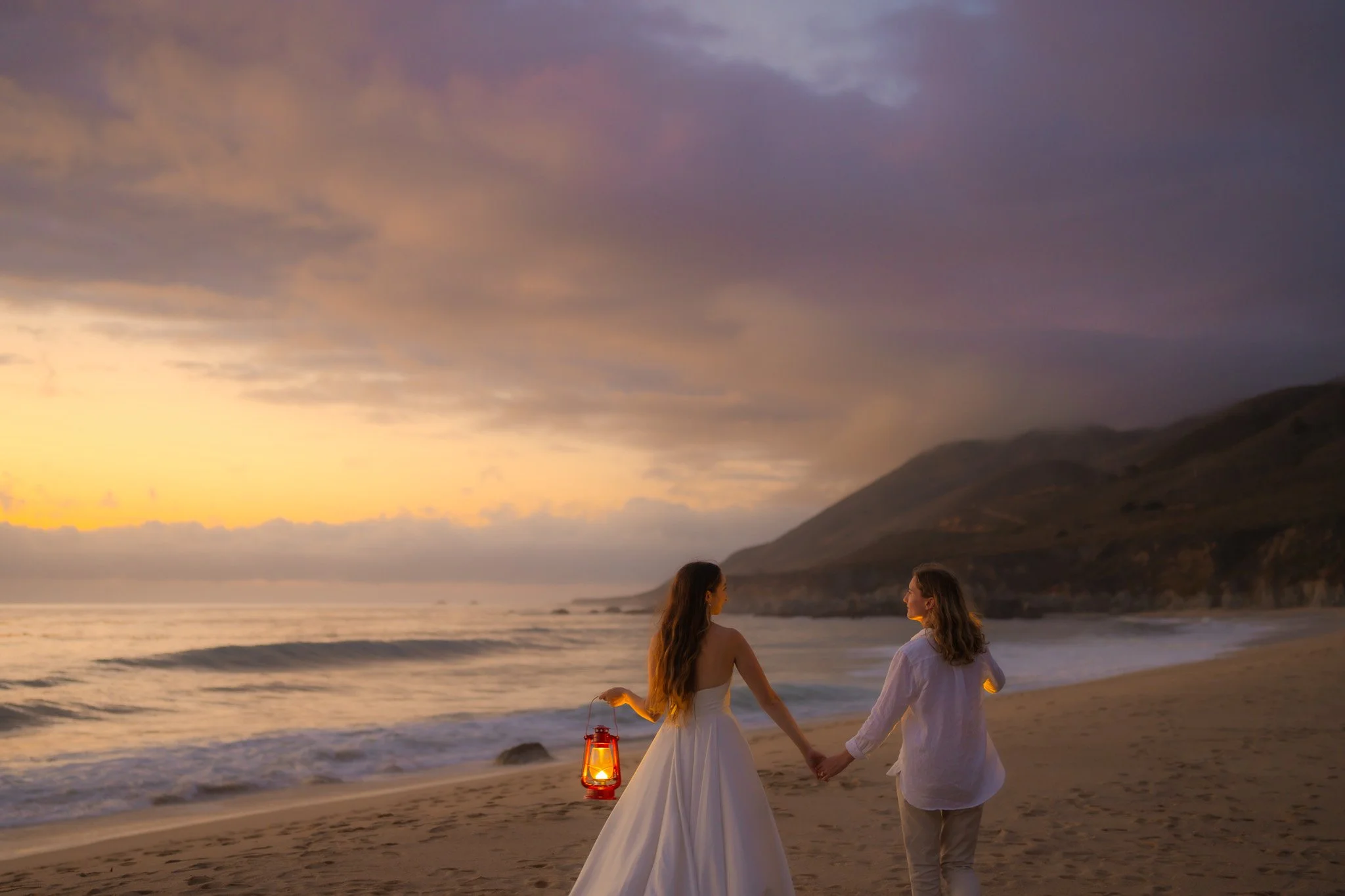 Two women holding hands walking on the beach at sunset with mountains in the background.