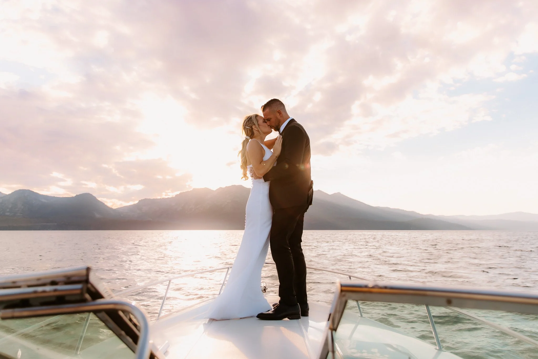 A couple dressed in wedding attire sharing a kiss on a boat at sunset with mountains in the background.