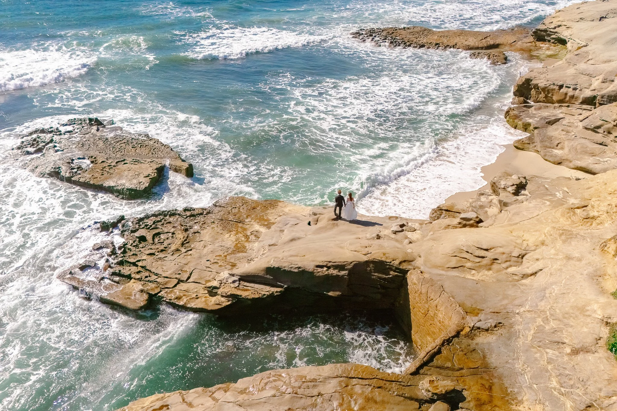 Two people in formal attire walk along a rocky coastal area beside the ocean.