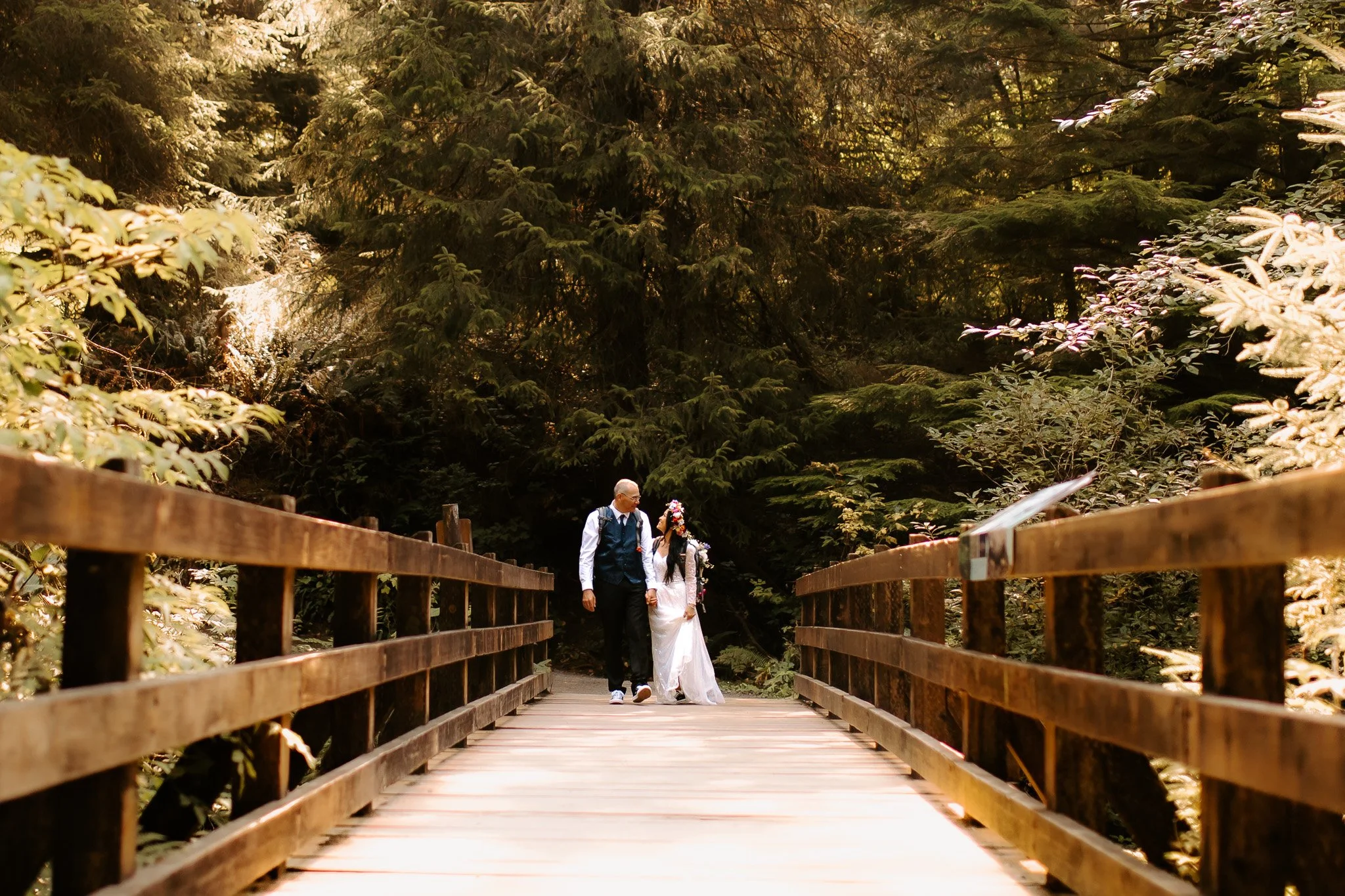 A bride and groom walking hand in hand on a wooden bridge surrounded by lush green trees.
