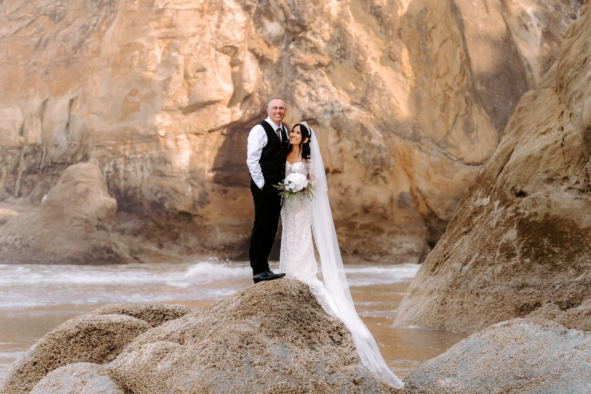A bride and groom standing on rocks near the ocean, with a rocky cliff in the background, dressed in wedding attire and smiling.