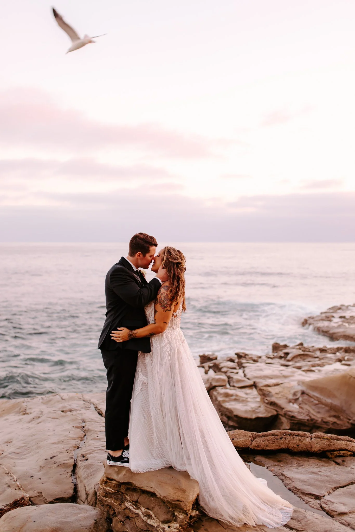A couple dressed in wedding attire standing and kissing on rocks by the ocean at sunset