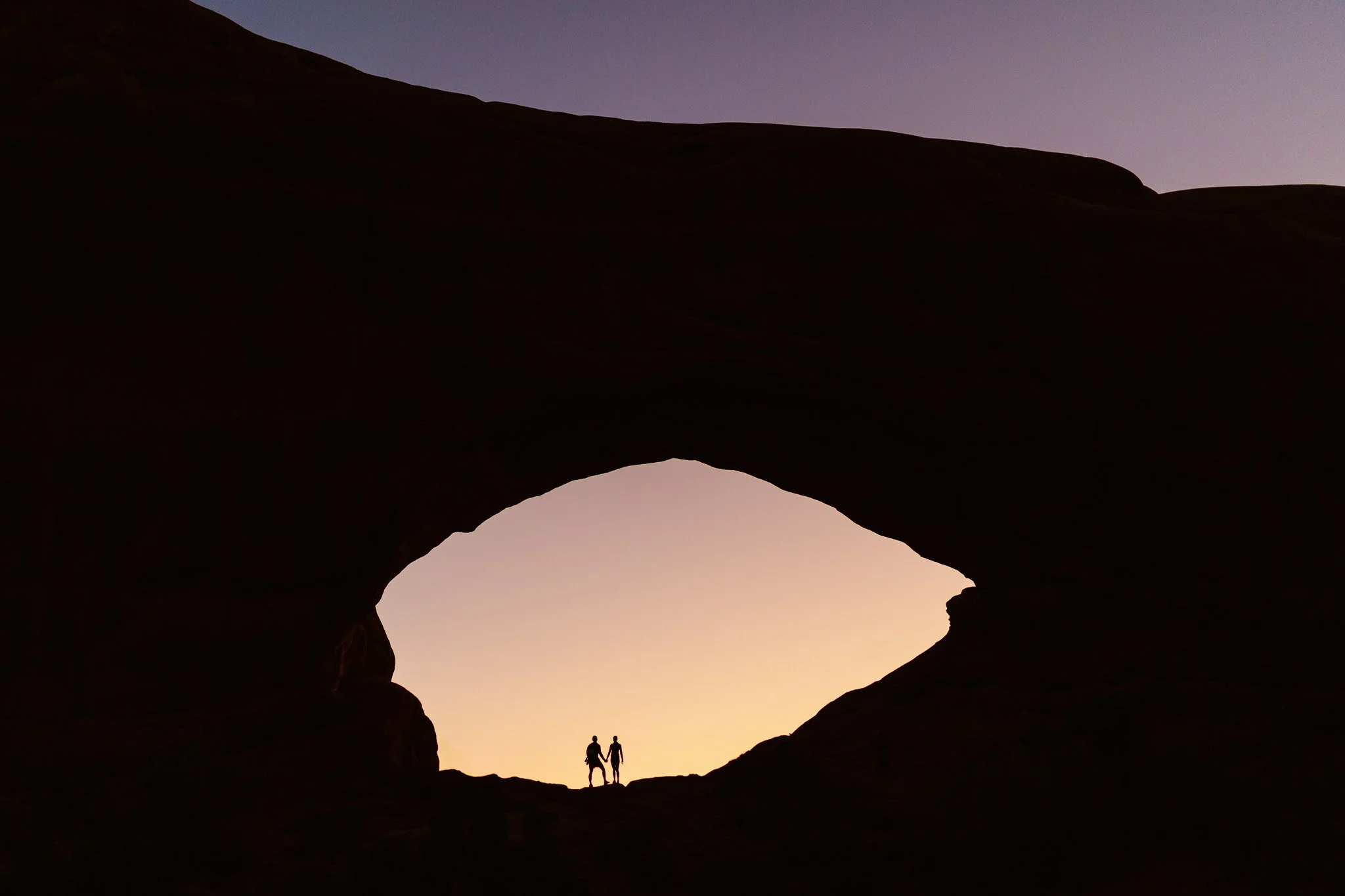 Silhouettes of two people walking through a canyon at sunset, with a large arch framing the sky in the background.