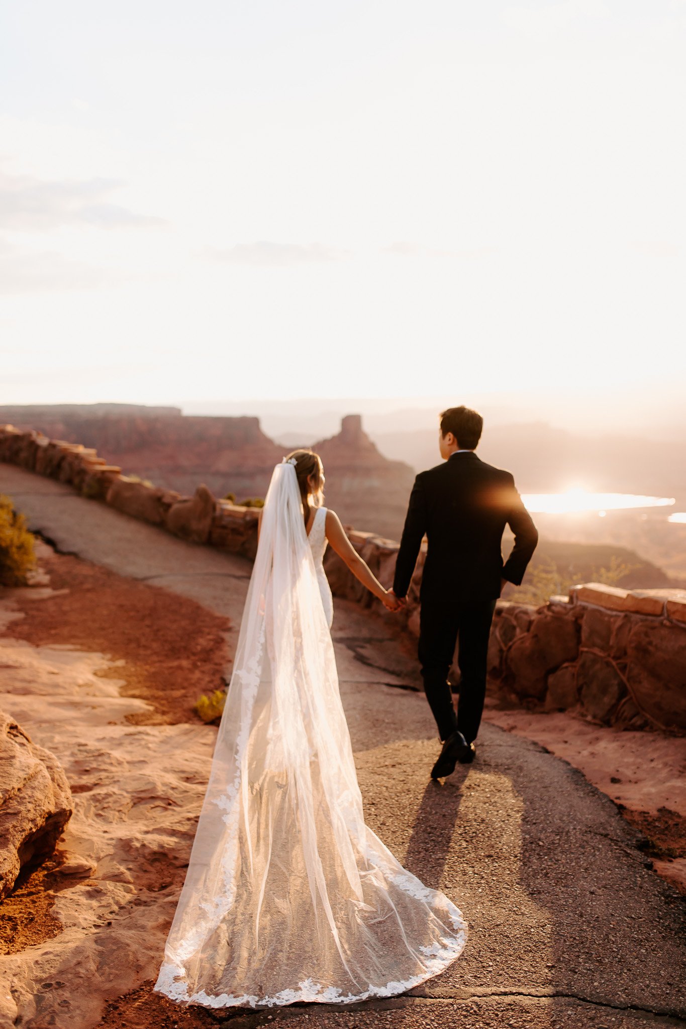 A bride and groom walking hand in hand on a scenic overlook at sunset, with the Grand Canyon in the background.
