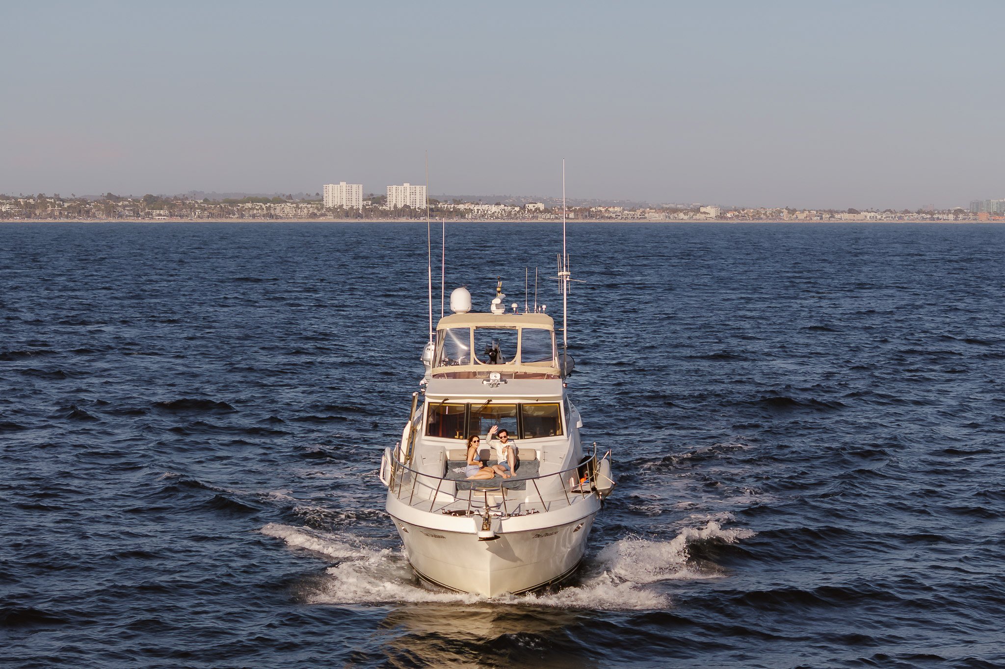 A white yacht cruising on the ocean with two people sitting on the front deck, raising their hands, and a city skyline with high-rise buildings in the background.