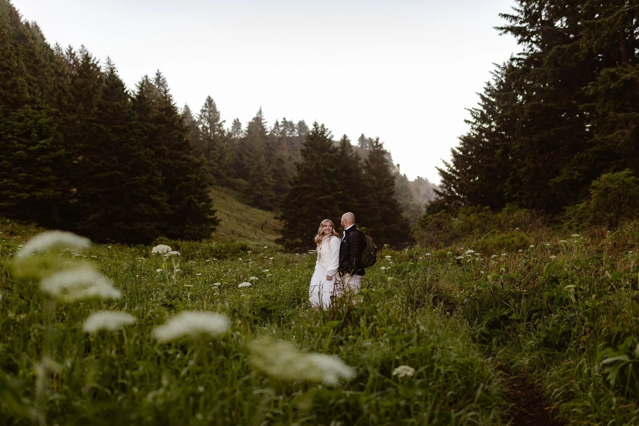 A couple standing in a field of wildflowers surrounded by tall evergreen trees under a partly cloudy sky.