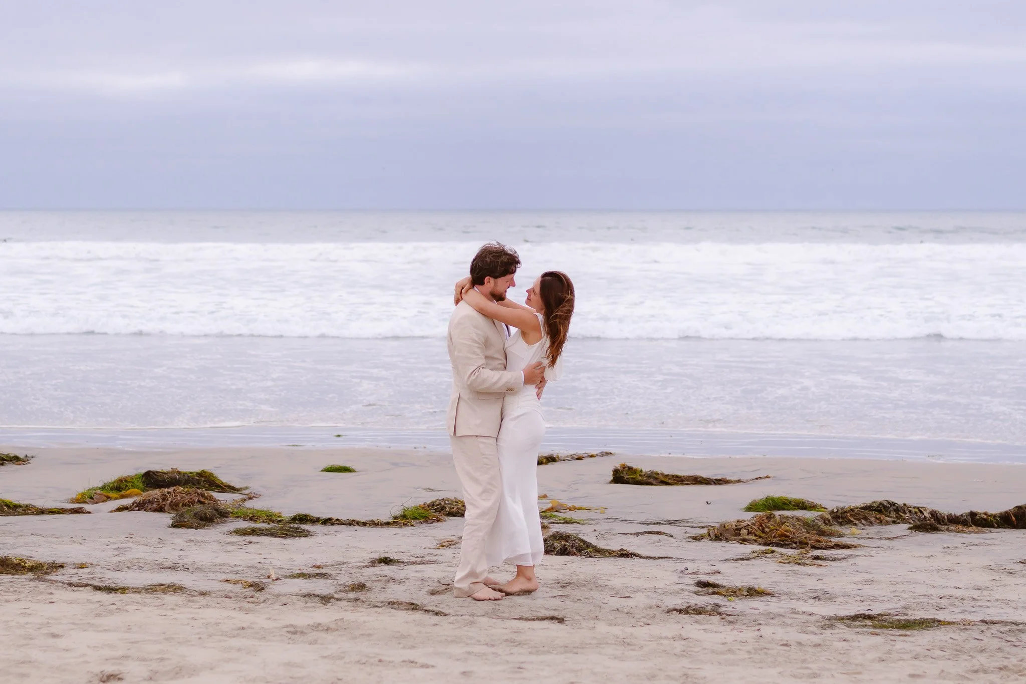 A couple embracing on a beach during overcast weather, standing close with heads touching, the man in a beige suit and the woman in a white dress, with ocean waves and cloudy sky in the background.