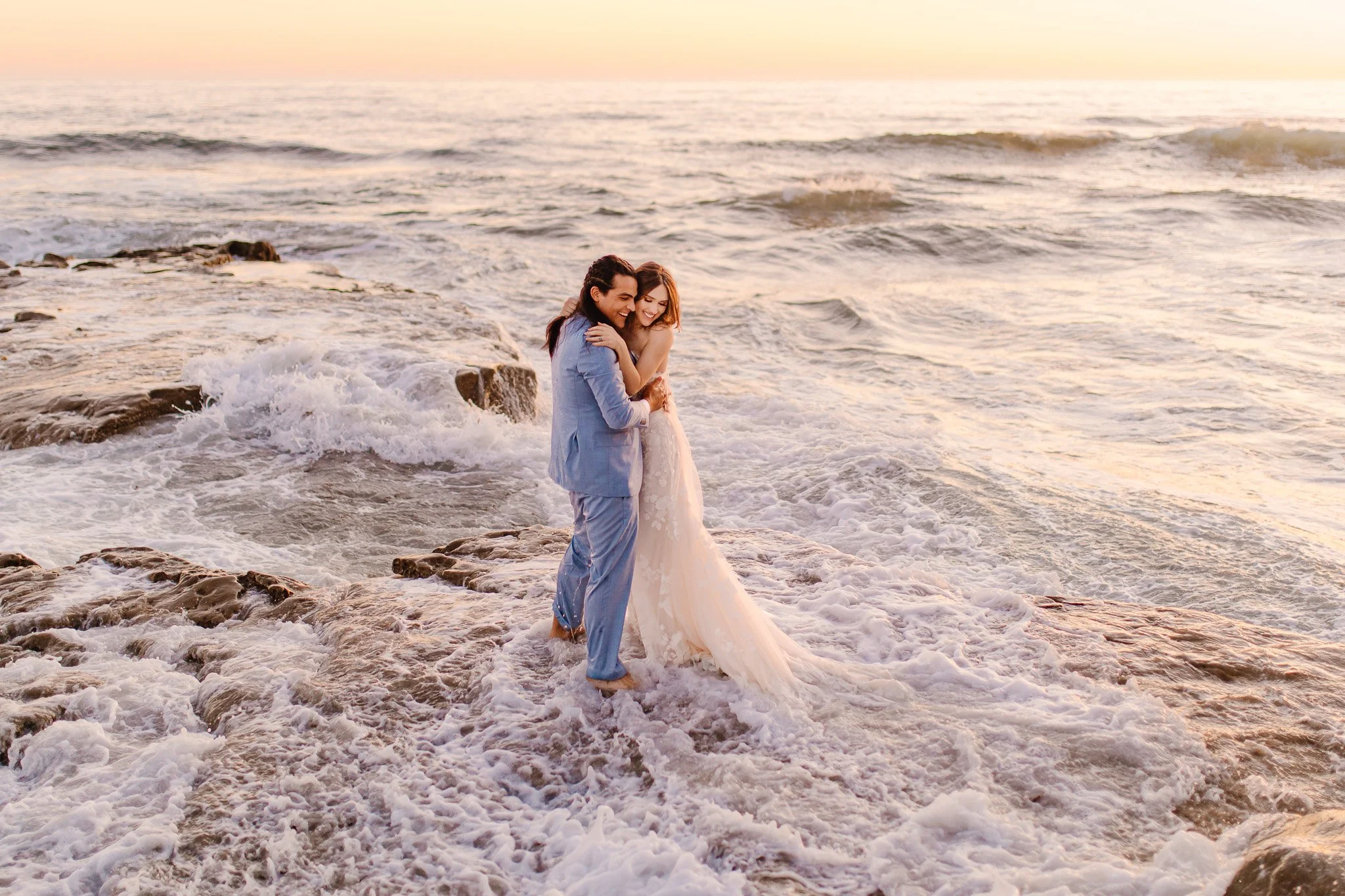 A couple in wedding attire standing on the seashore with waves crashing around them, during sunset.