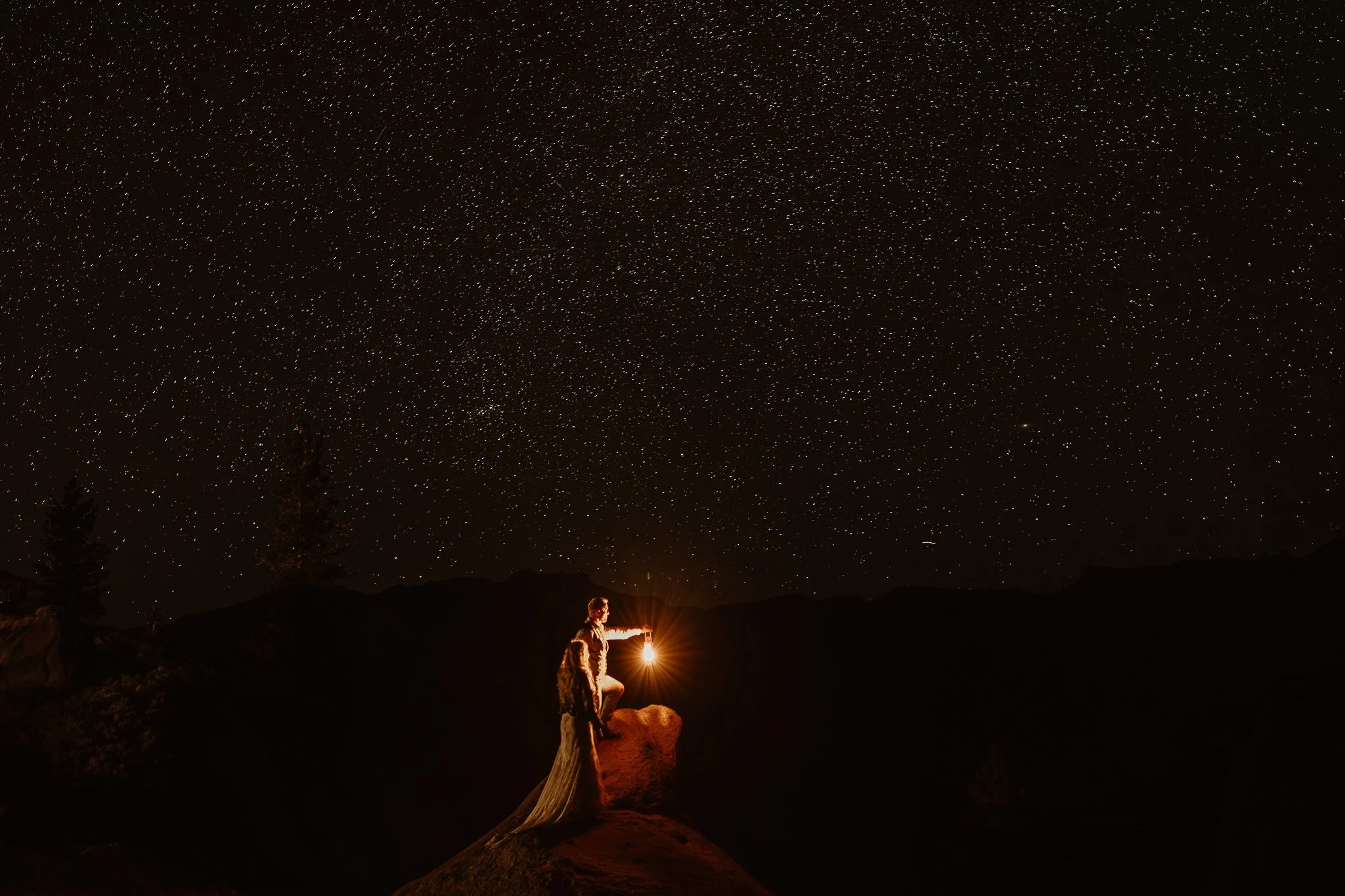 A person holding a lantern while standing on rocks under a starry night sky with mountains in the distance.