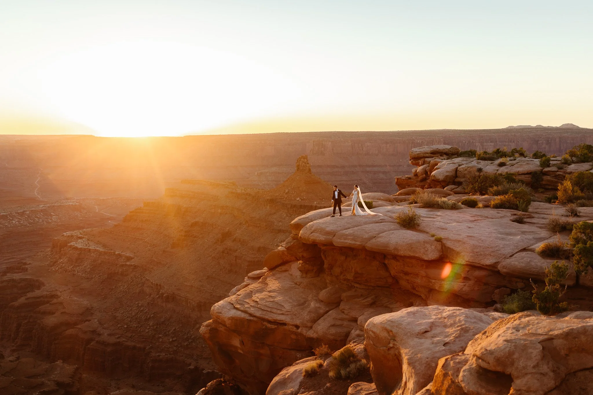 A couple in wedding attire standing on a rocky ledge in a desert canyon at sunset, holding hands as the sun sets behind the canyon walls.