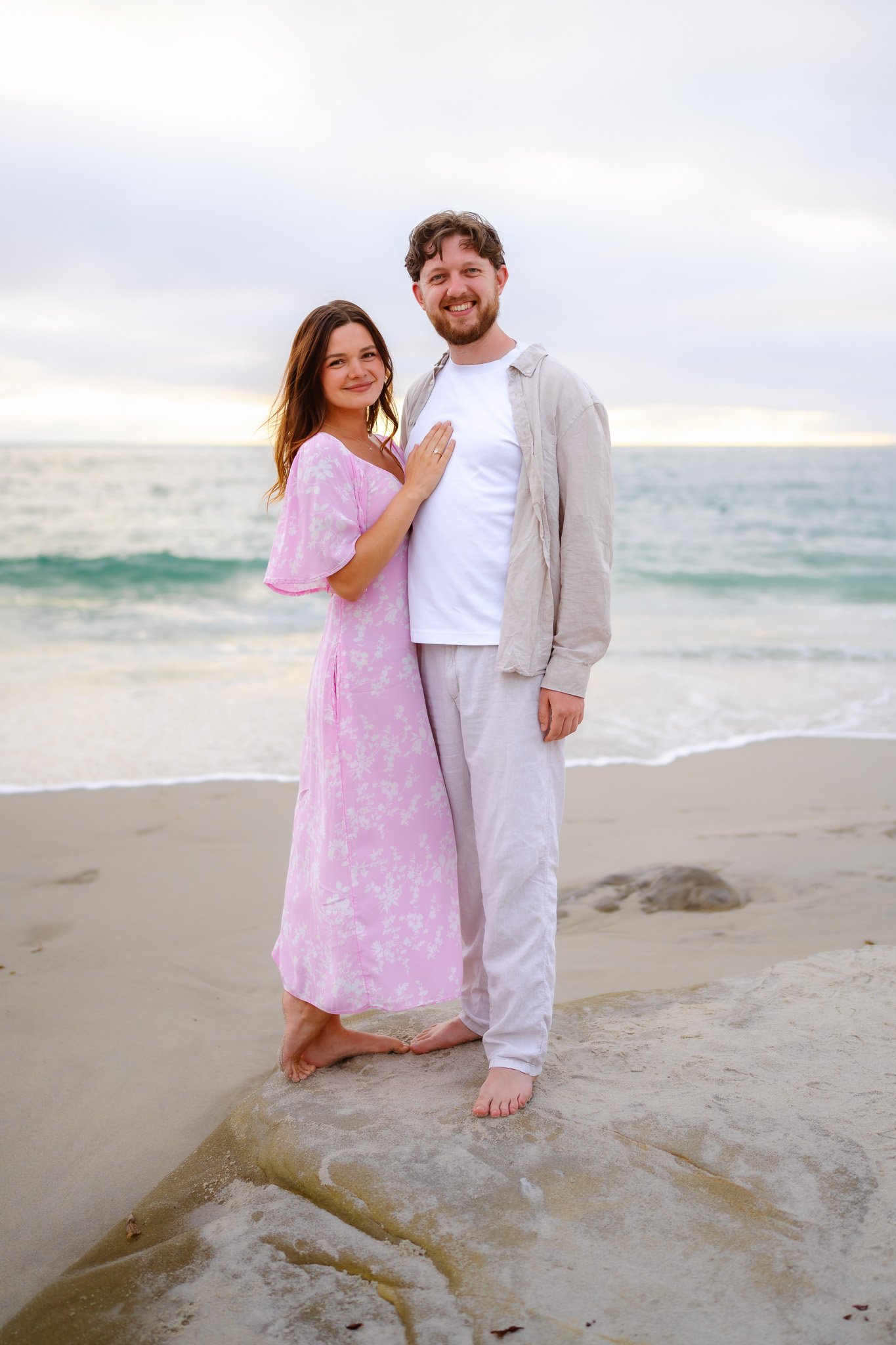 A smiling young woman in a pink floral dress and a smiling young man in white clothing standing on a beach with ocean waves behind them. They are barefoot, with one hand on the man's chest, close to each other, near the water and rocks.