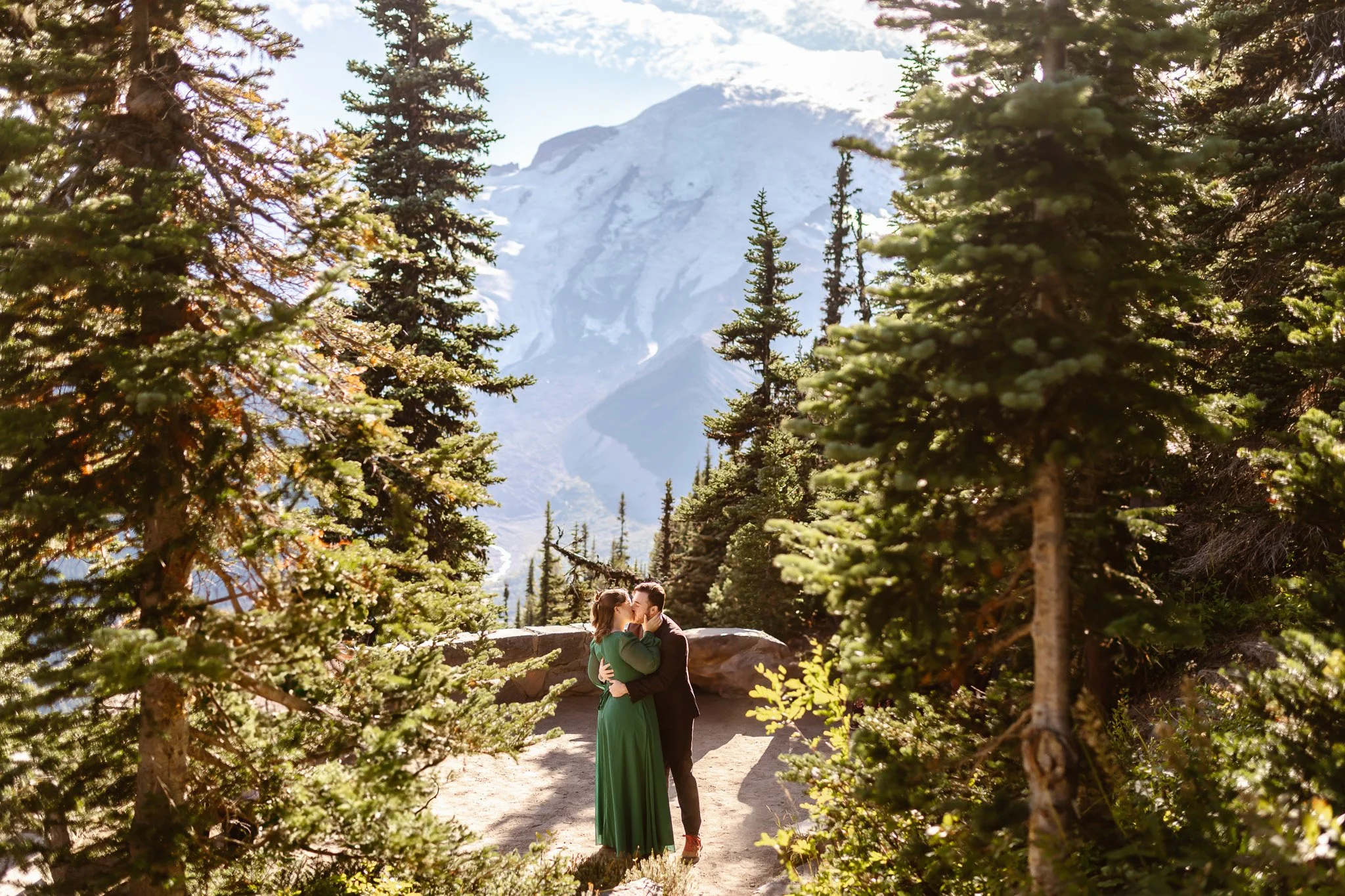 A couple kissing outdoors in a forested mountain area, surrounded by tall evergreen trees with a snow-capped mountain in the background.
