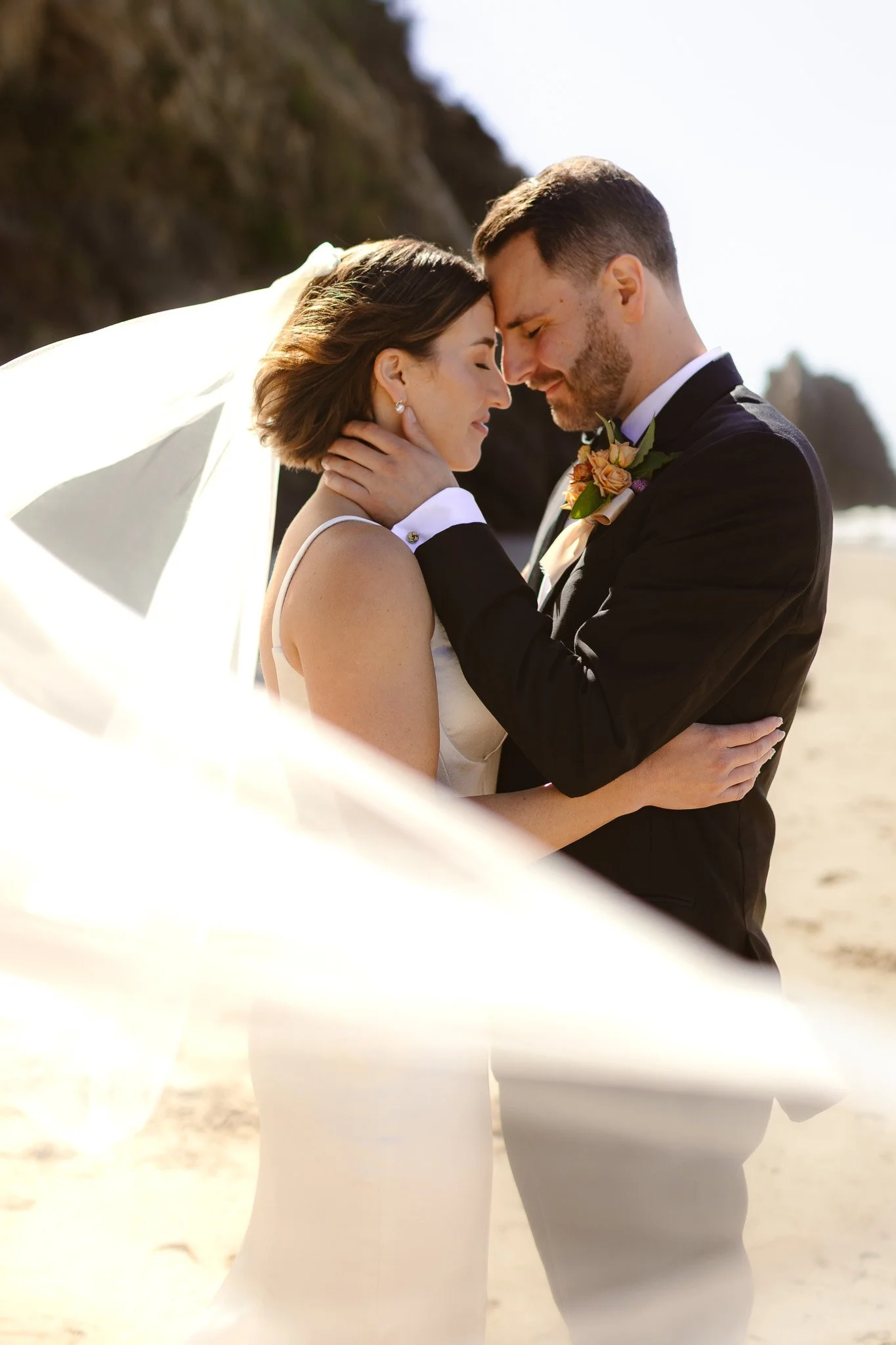 A bride and groom on the beach, touching foreheads and holding each other during their wedding.