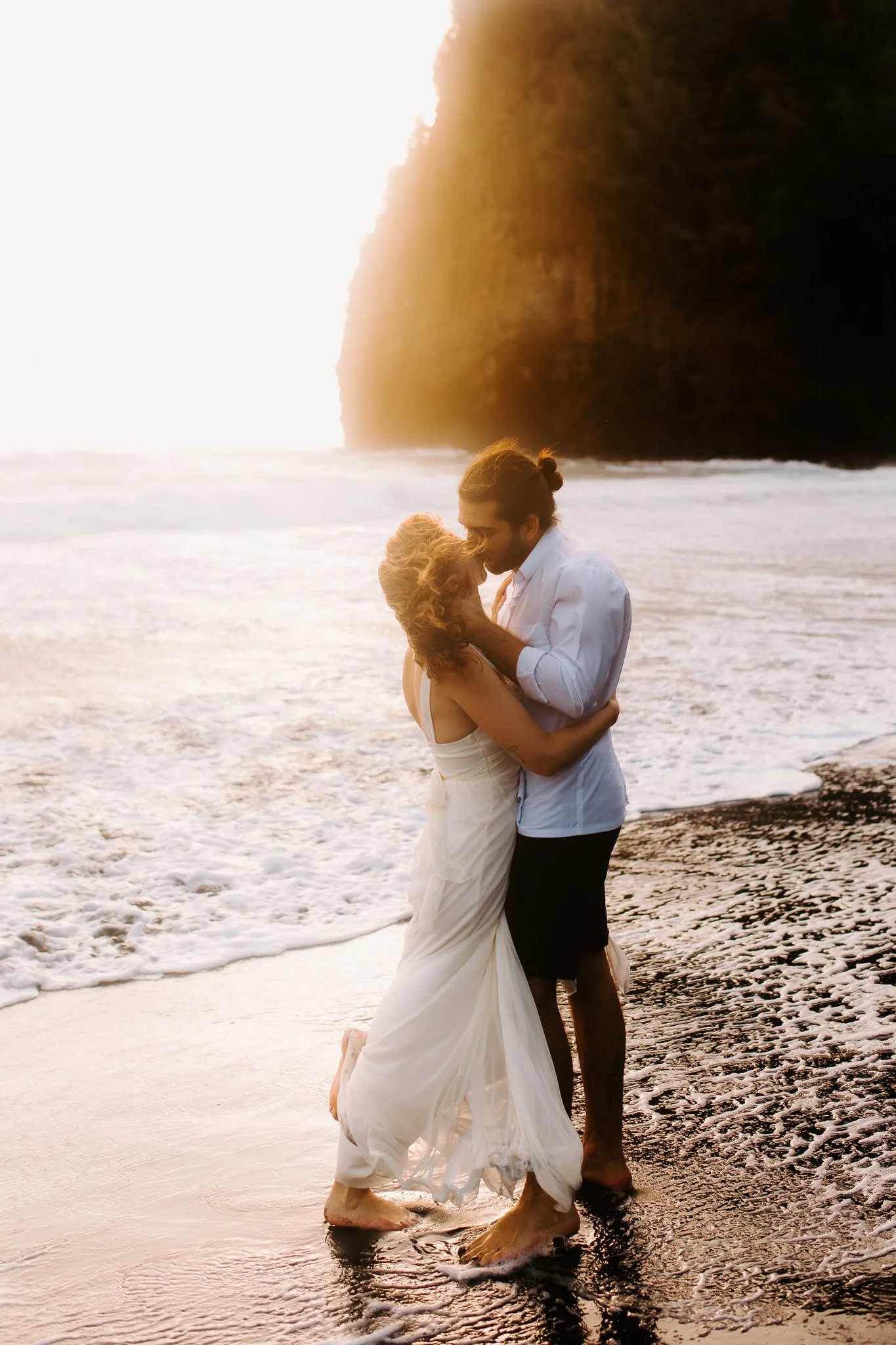 A couple kissing on a beach during sunset, with waves and a cliff in the background.