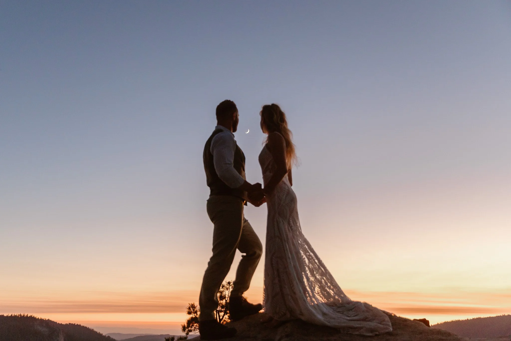 A couple stands on a rocky hilltop holding hands and looking into each other's eyes during sunset. The woman is wearing a white dress with a long train, and the man is wearing a vest and trousers. The sky is clear with a waxing crescent moon visible 