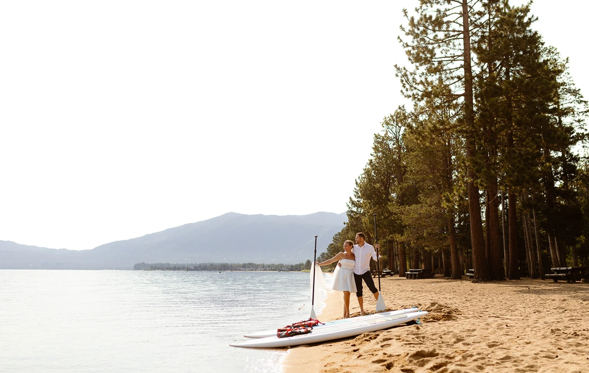 A couple stands on the beach near two stand-up paddleboards, with a lake and mountains in the background, and tall trees lining the shoreline.