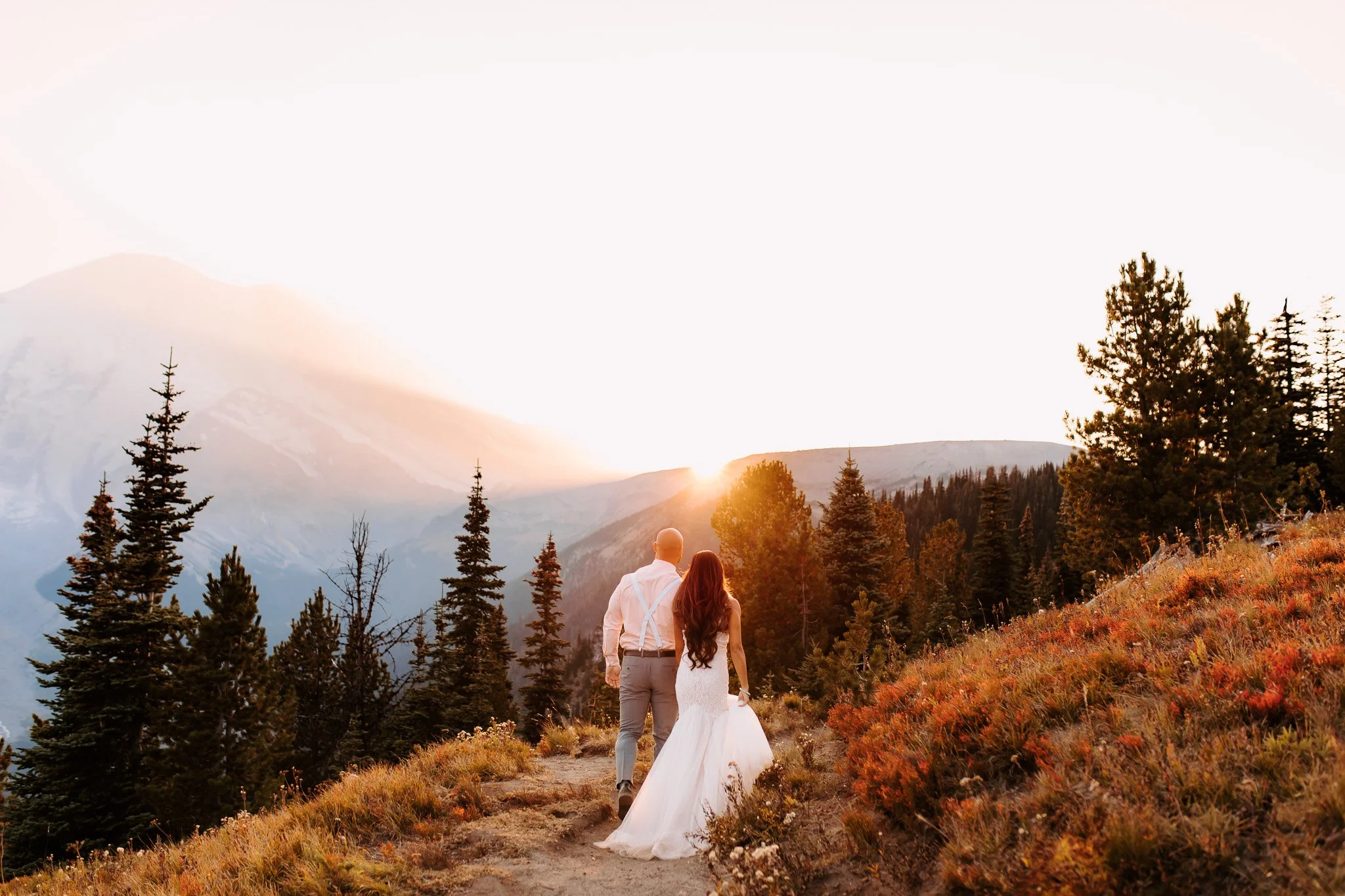 A couple dressed in wedding attire walk along a mountain trail at sunset, surrounded by trees and overlooking distant mountains and a body of water.