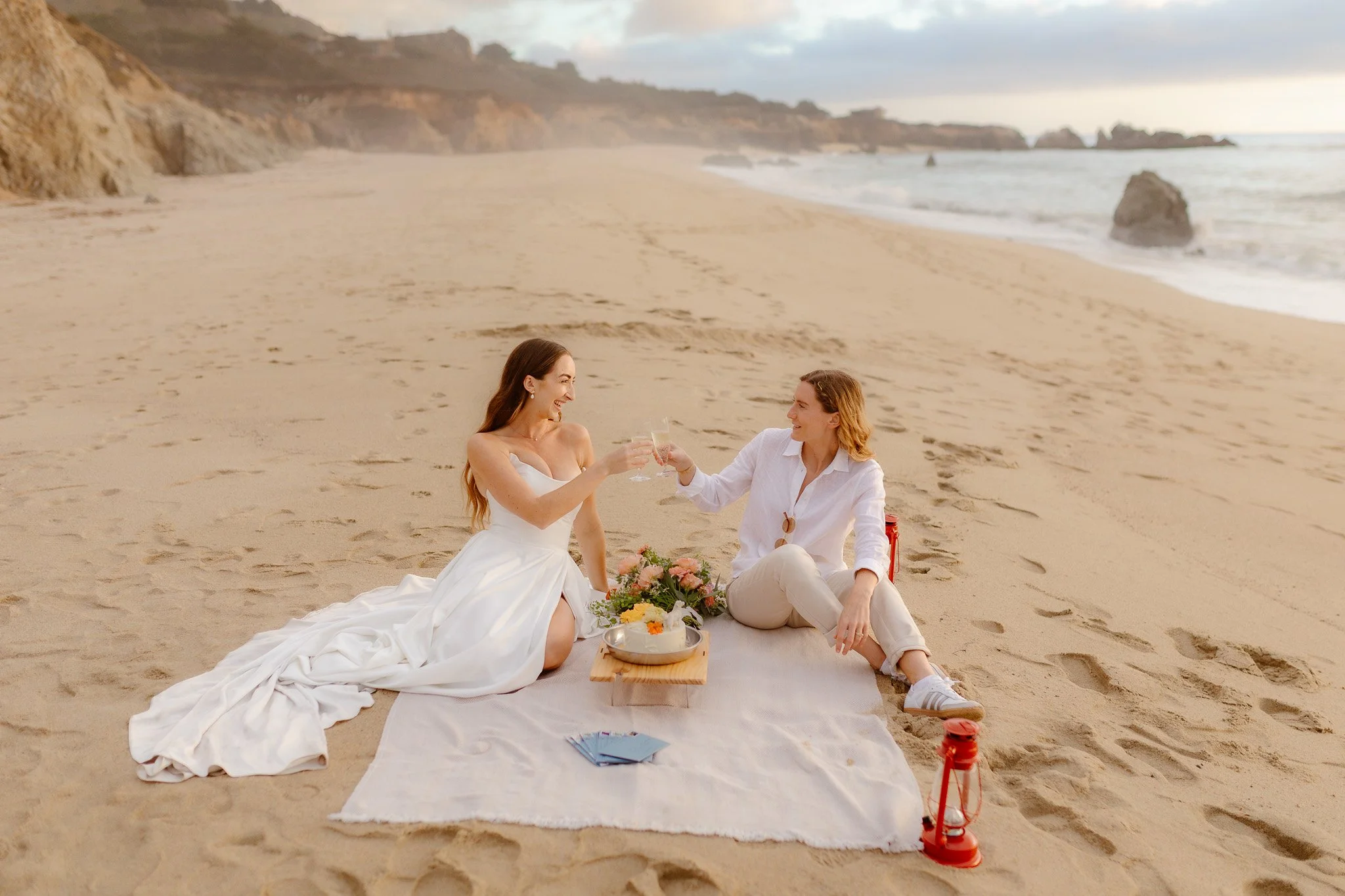 Two women in wedding attire sitting on a blanket at the beach, toasting with champagne glasses, surrounded by flowers, with the ocean and cliffs in the background.