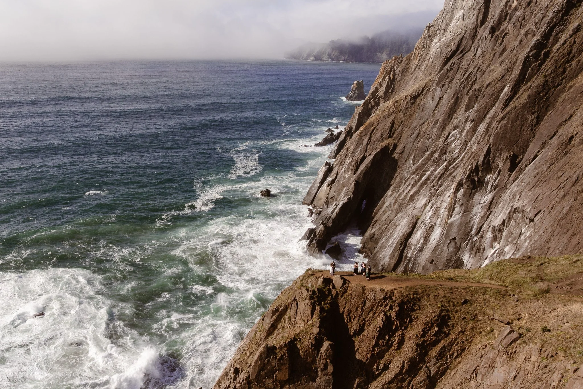 View of the ocean crashing against rocky cliffs, with a group of people standing at the base of the cliff.