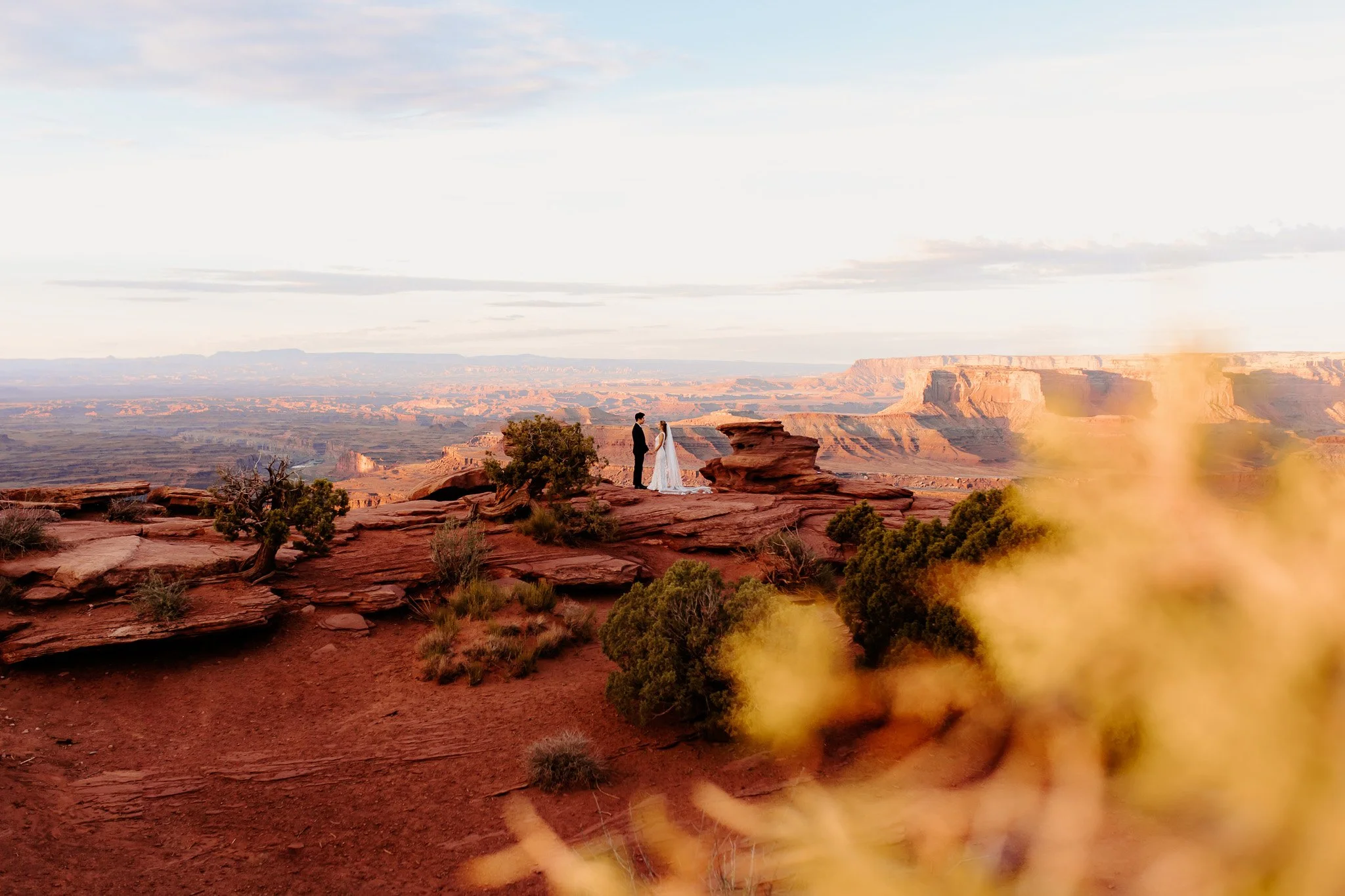A couple dressed in wedding attire stands facing each other on a rocky ledge in the Grand Canyon at sunset, with expansive canyon views and a cloudy sky in the background.